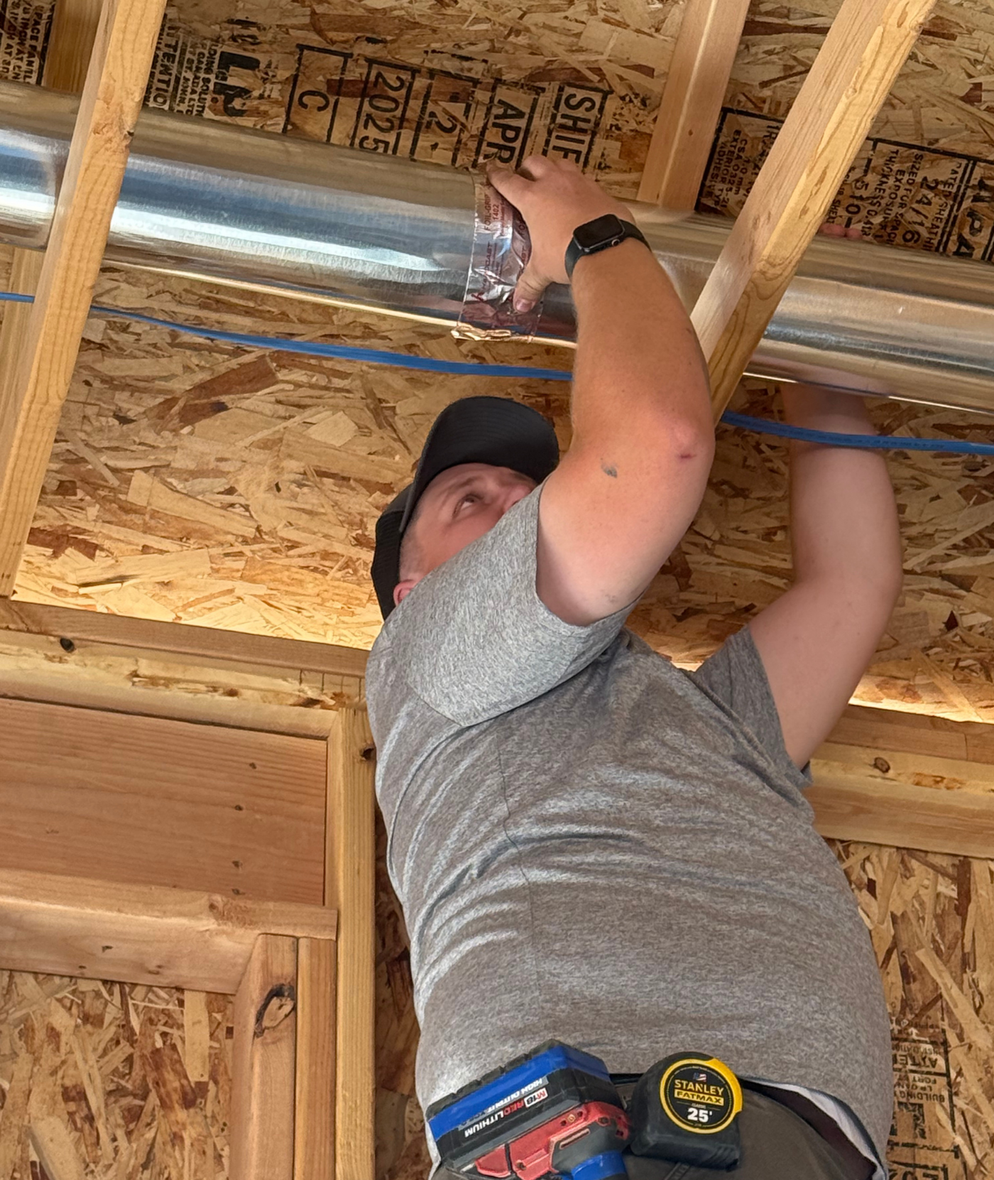 Man in a gray shirt installs ductwork in a wooden ceiling.