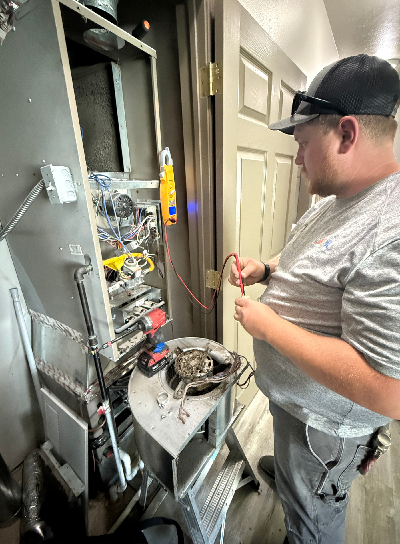 HVAC technician repairs unit, using multimeter. Outdoors, wearing hat and watch, working with wires.