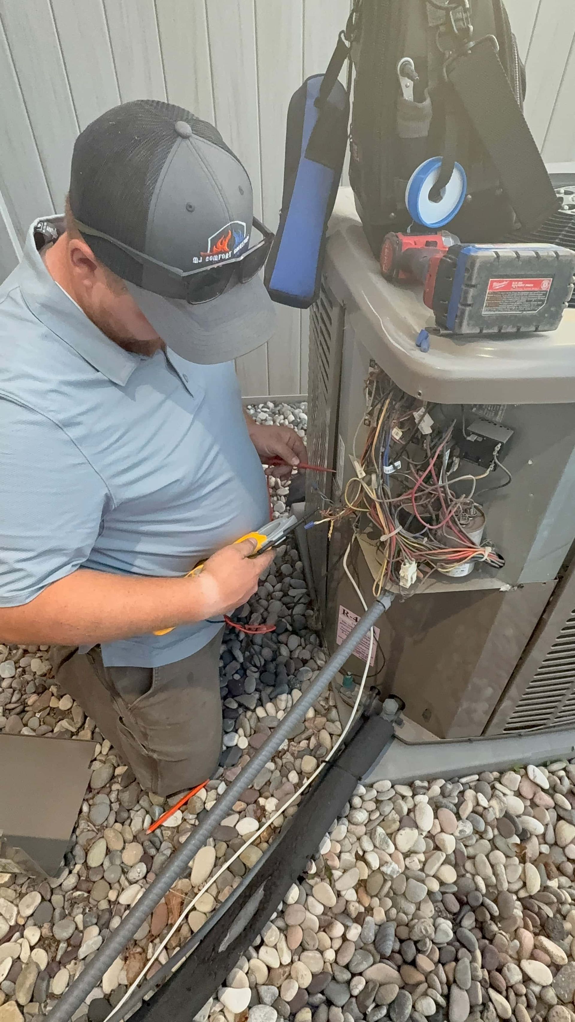 HVAC technician in a cap working on outdoor AC unit wiring near a fence.