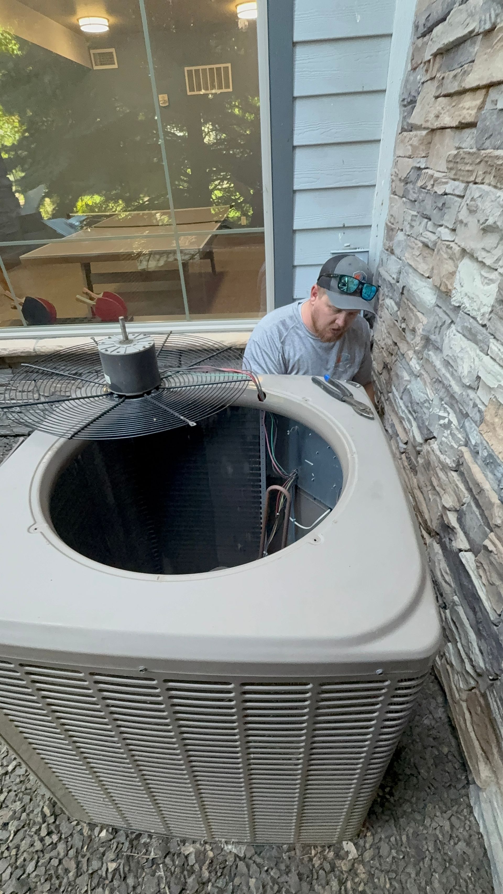 A man in a hat inspects an AC unit outside a building with stone siding.