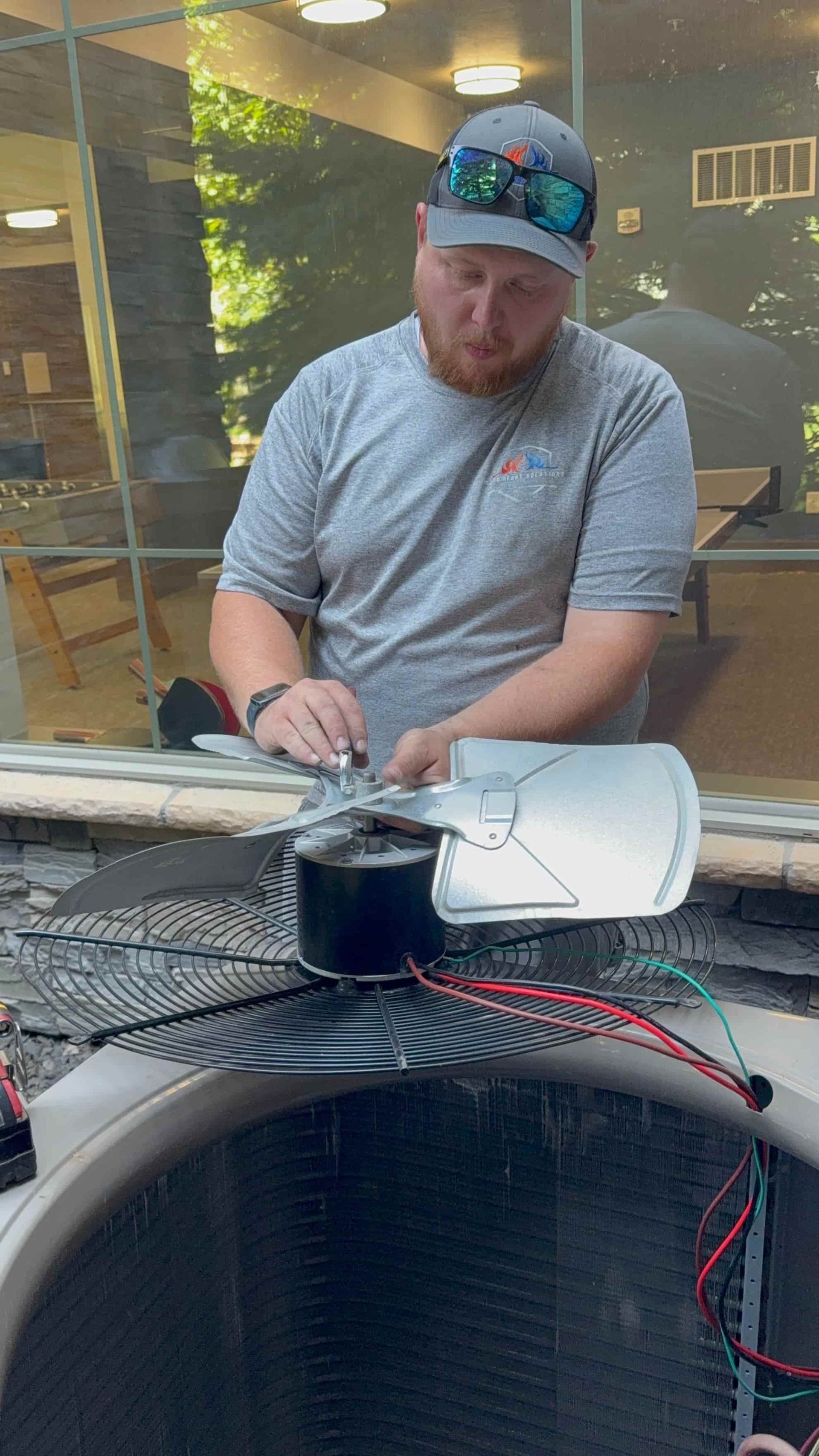 A man in a grey shirt is working on a fan, likely for an air conditioner, outdoors.