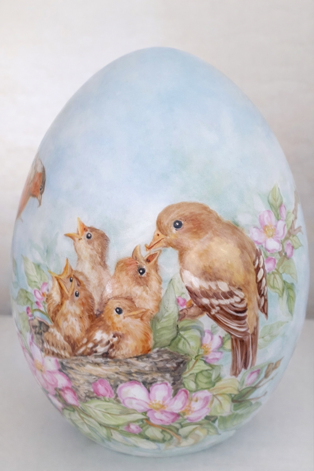 Uovo dipinto con uccelli che nutrono i pulcini in un nido, sfondo di cielo azzurro tenue, fiori rosa.