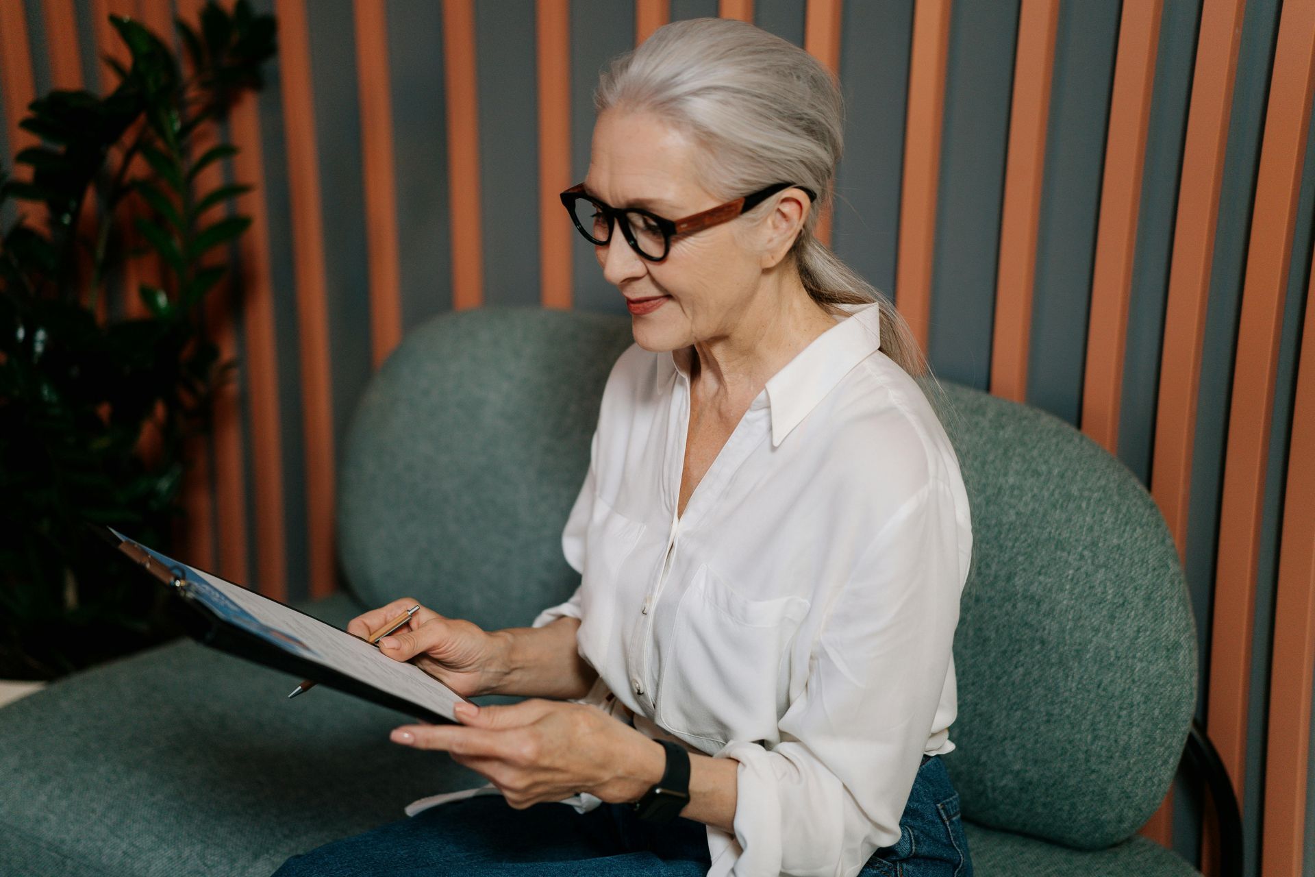 An elderly woman is sitting on a couch reading a book.