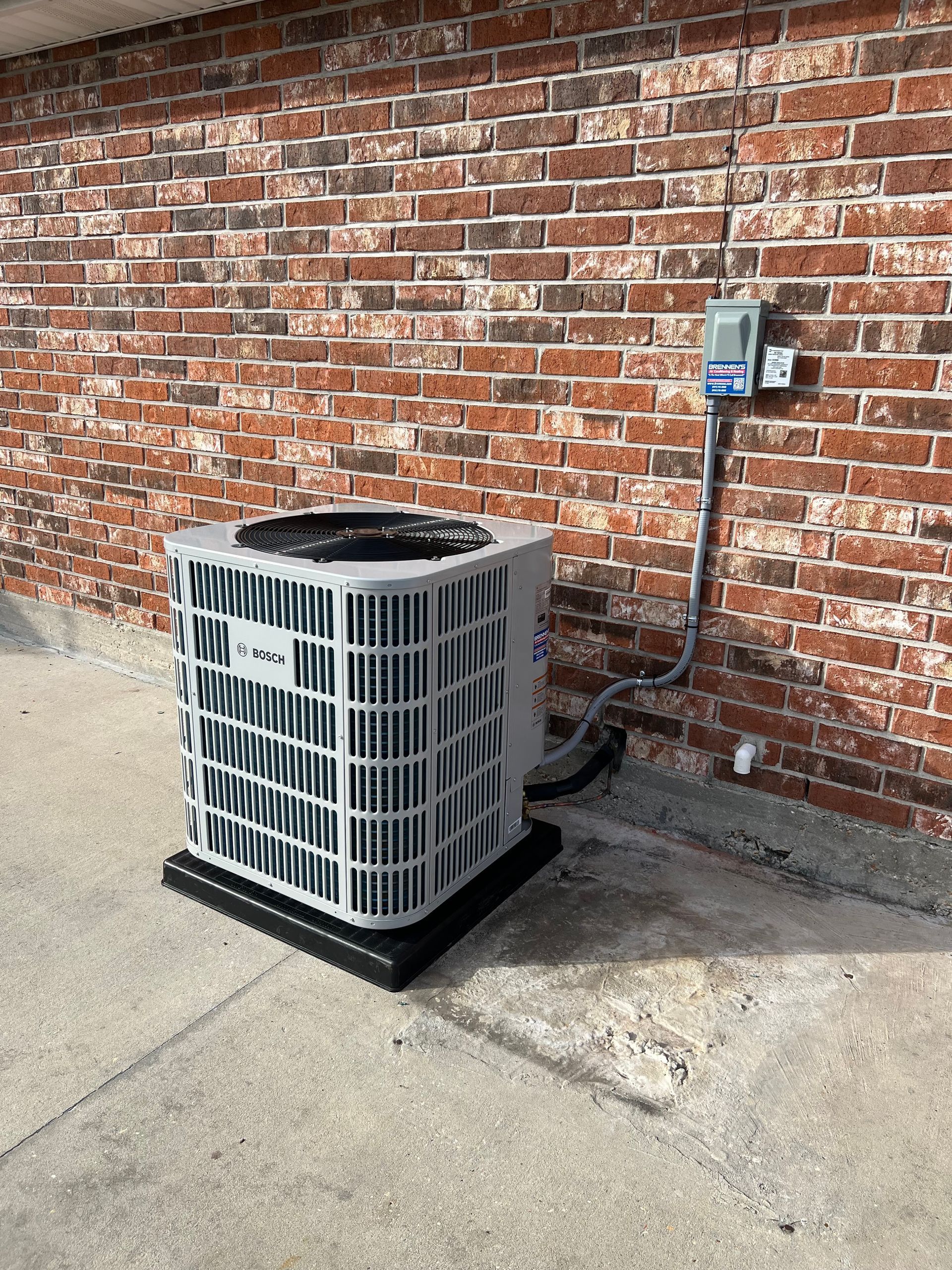 An air conditioner is sitting on the sidewalk in front of a brick wall.