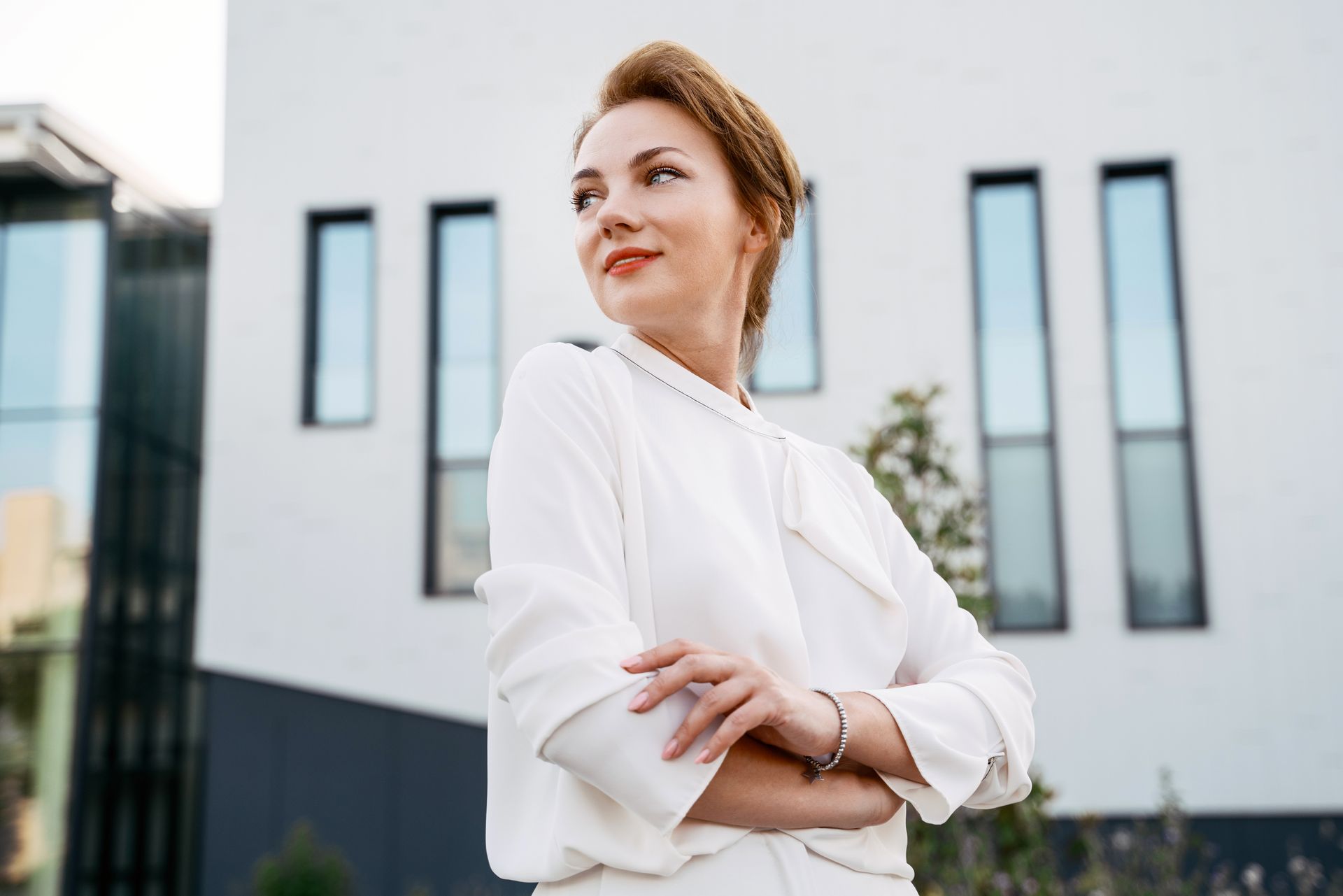 A woman in a white shirt is standing in front of a building with her arms crossed.