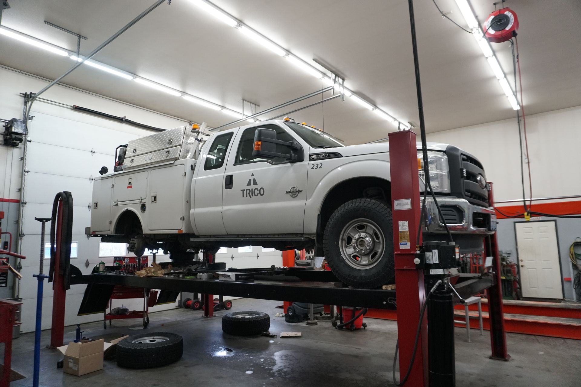 White truck on a lift in a garage. Interior shot with open bay door. Tools on the floor.