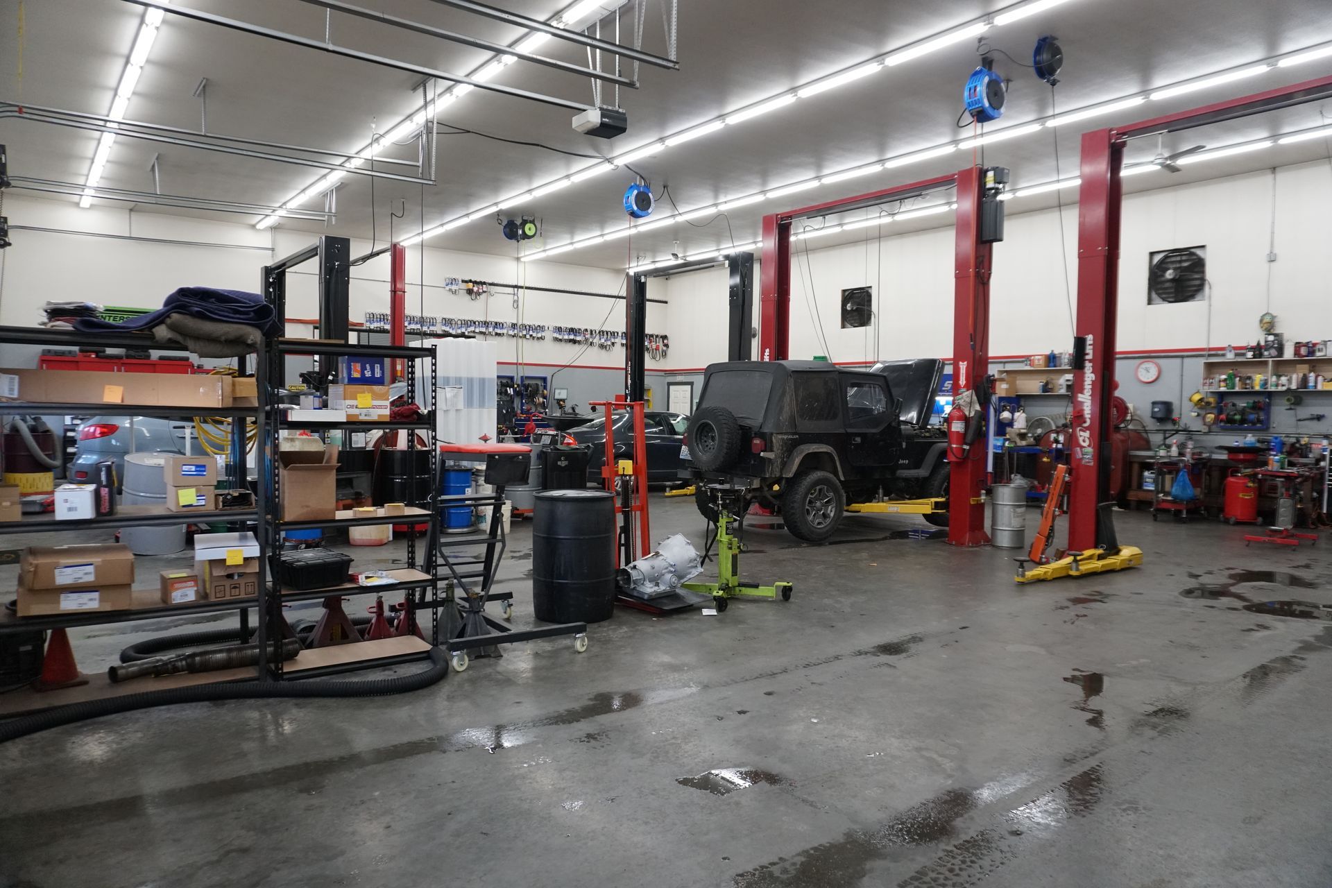 Inside a car repair shop. A black Jeep sits on a lift. Tools, equipment, and shelves line the space.