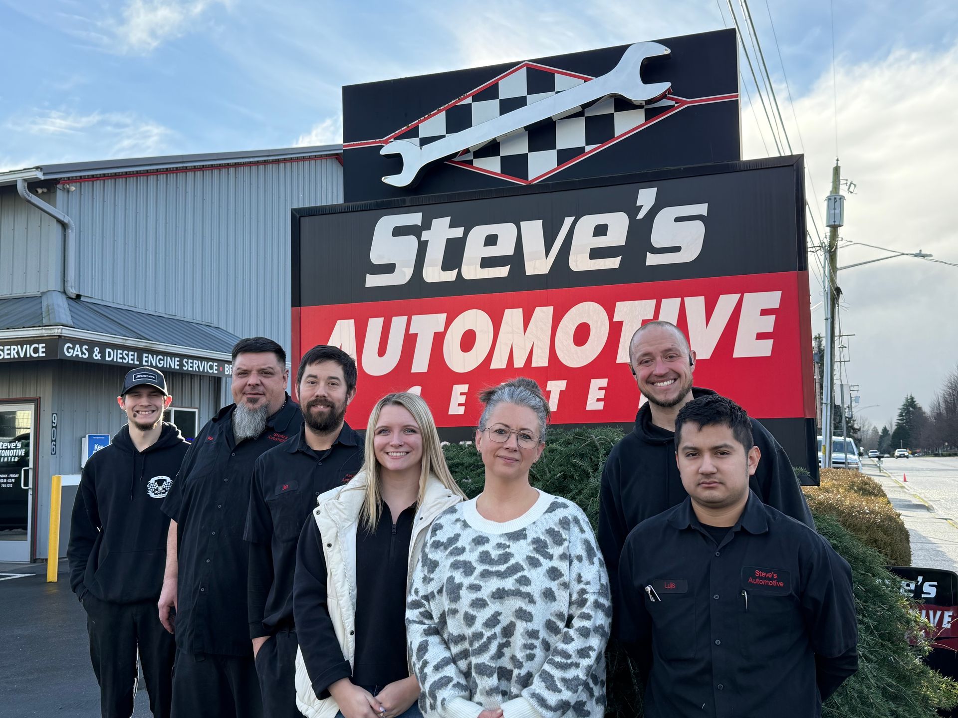 Employees posing in front of Steve's Automotive Center sign with checkered flag and wrench.