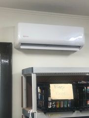 White air conditioner mounted on a wall above a shelf with black crates and a handwritten sign.