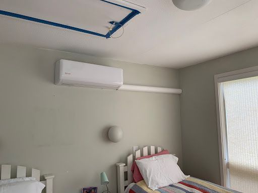 Air conditioner mounted on a pale gray wall in a bedroom, near a bed and window.