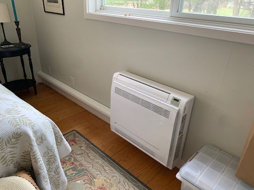 White wall-mounted heating unit next to a window and a small upholstered bench, with a bed visible on the left.