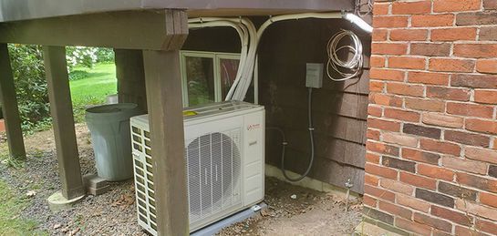 Outdoor air conditioning unit near a brick wall and under a wooden deck.