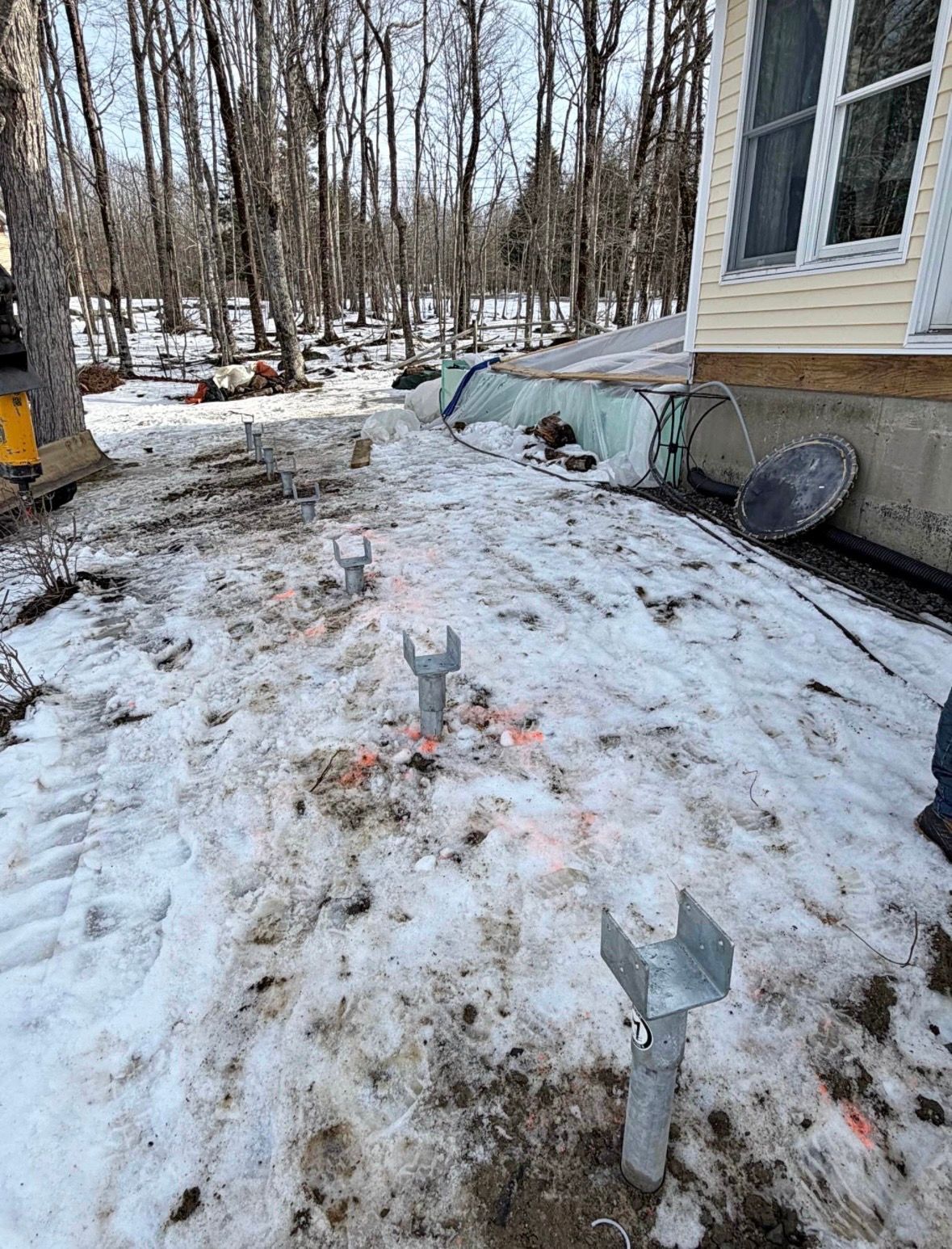 Metal deck post anchors installed in a snow-covered yard next to the concrete foundation of a house.