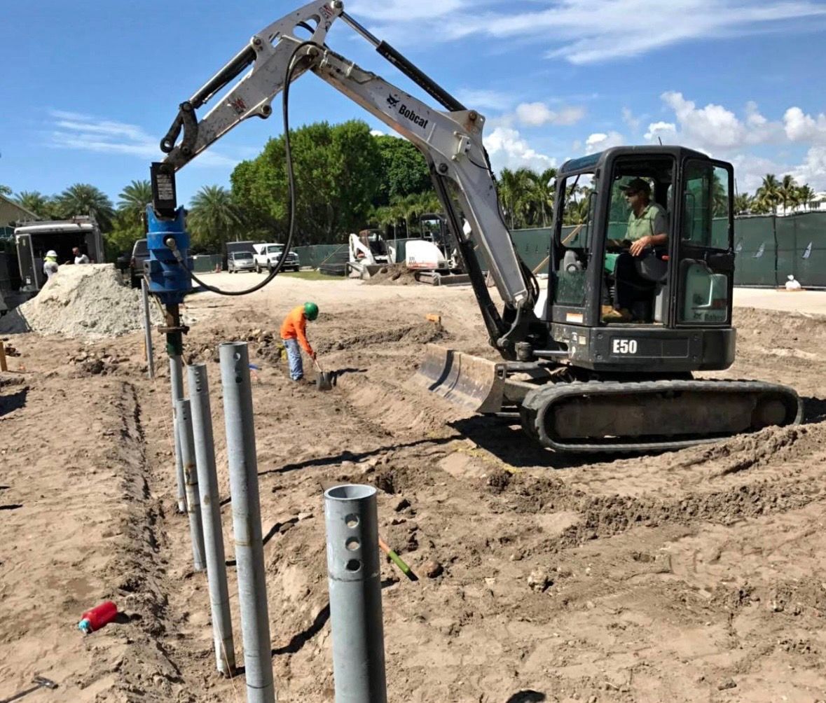 A Bobcat excavator drives metal posts into sandy ground at a construction site while a worker observes.