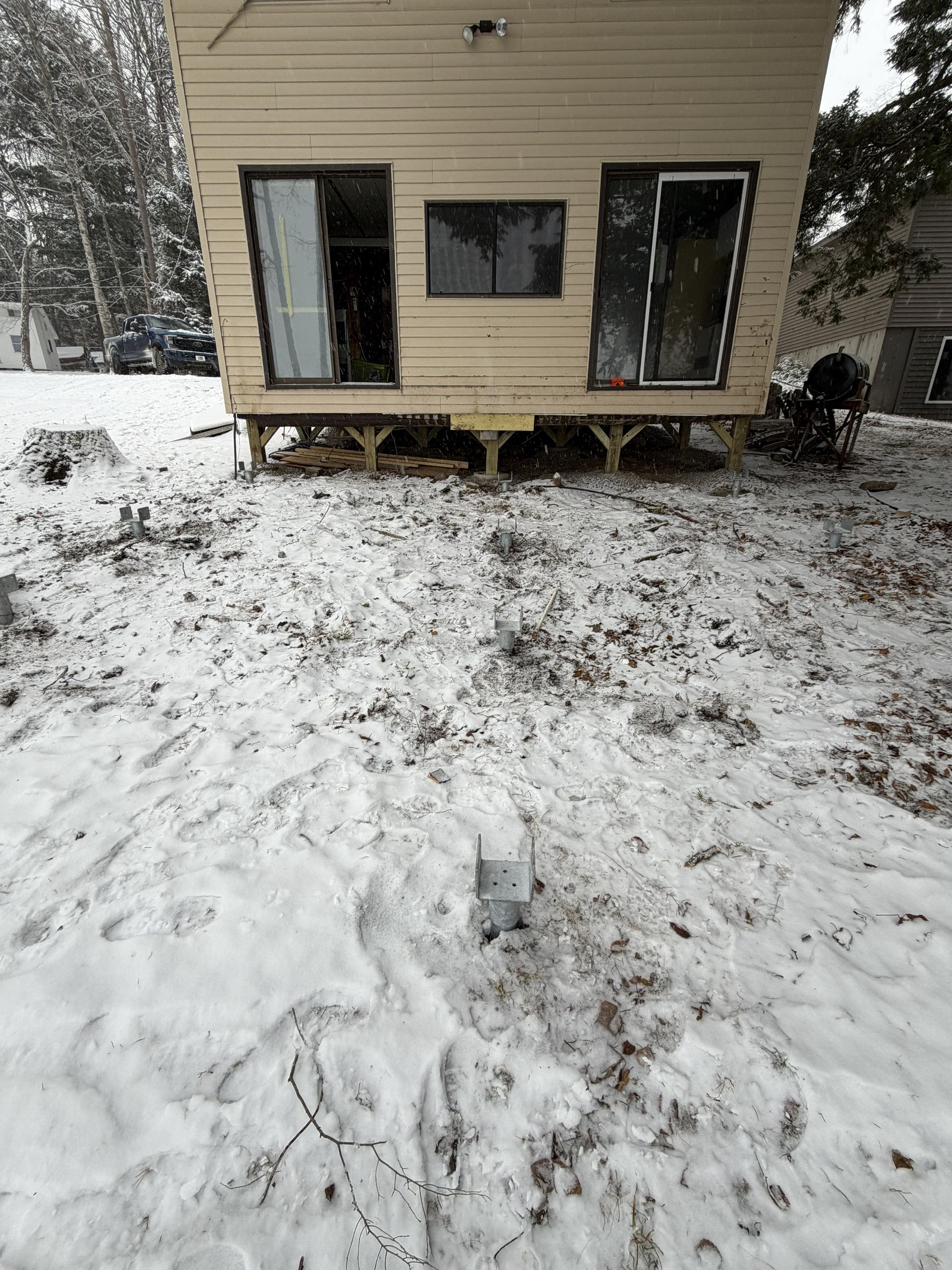 A light-colored mobile home on blocks in a snow-covered yard, with two doors flanking a central window.