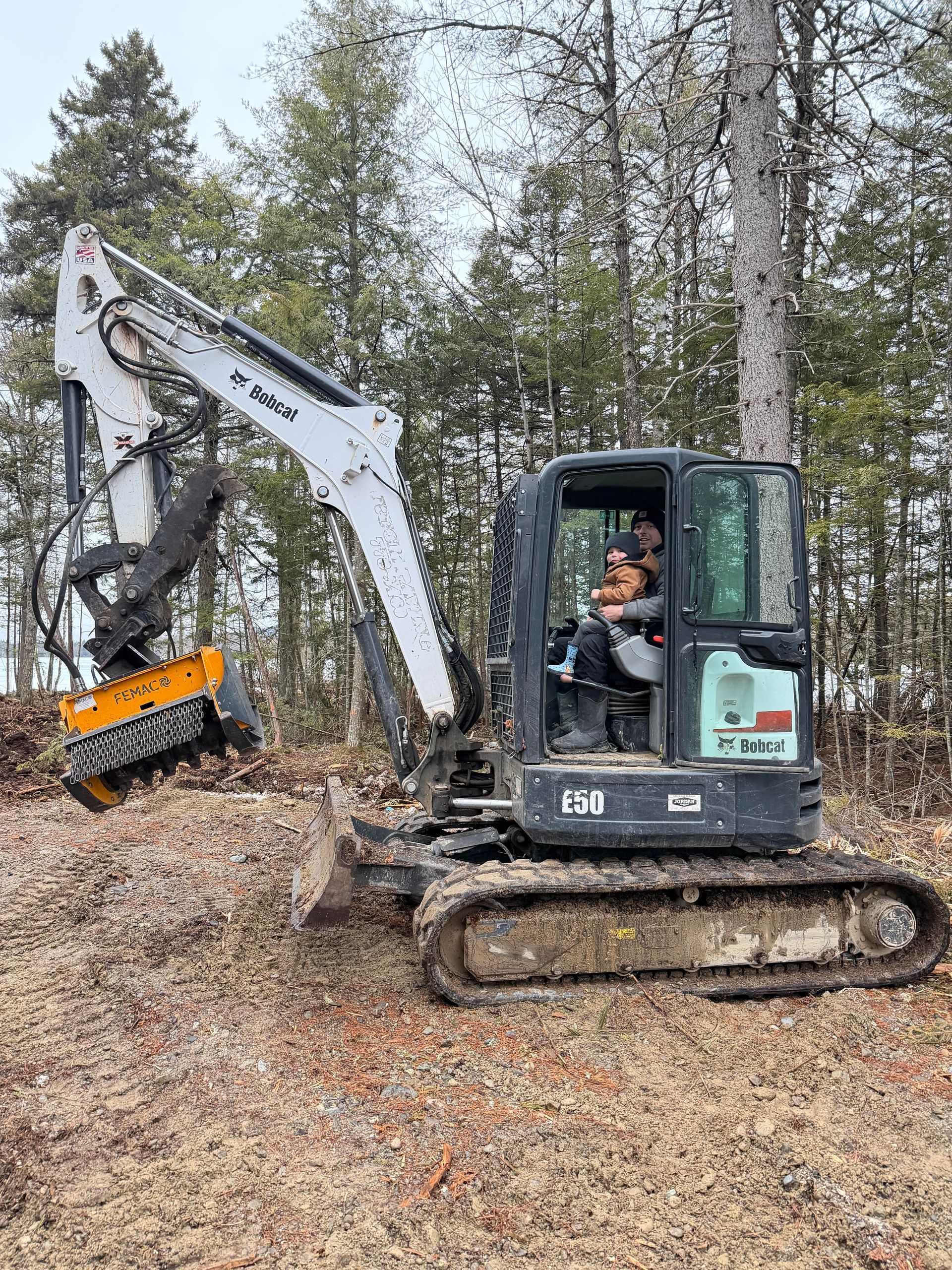 A person sits in the cab of a white and black Bobcat excavator equipped with a forestry mulcher in a wooded area.