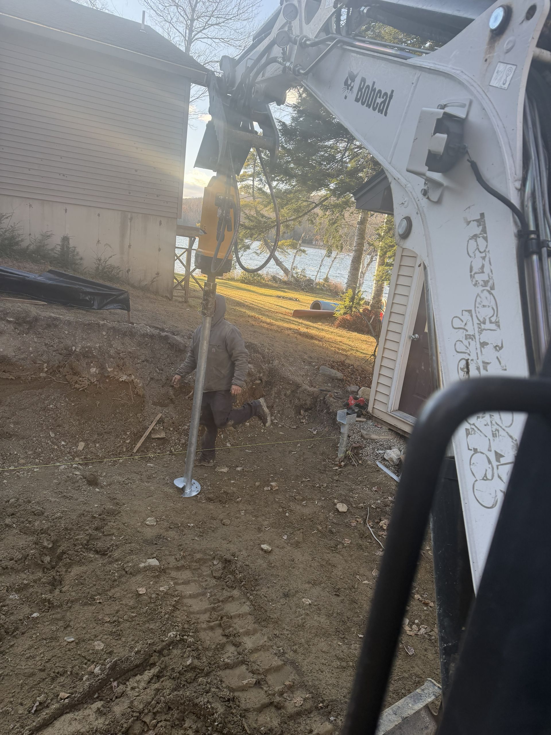 An excavator operates an auger attachment, drilling a metal pier into the ground at a construction site.