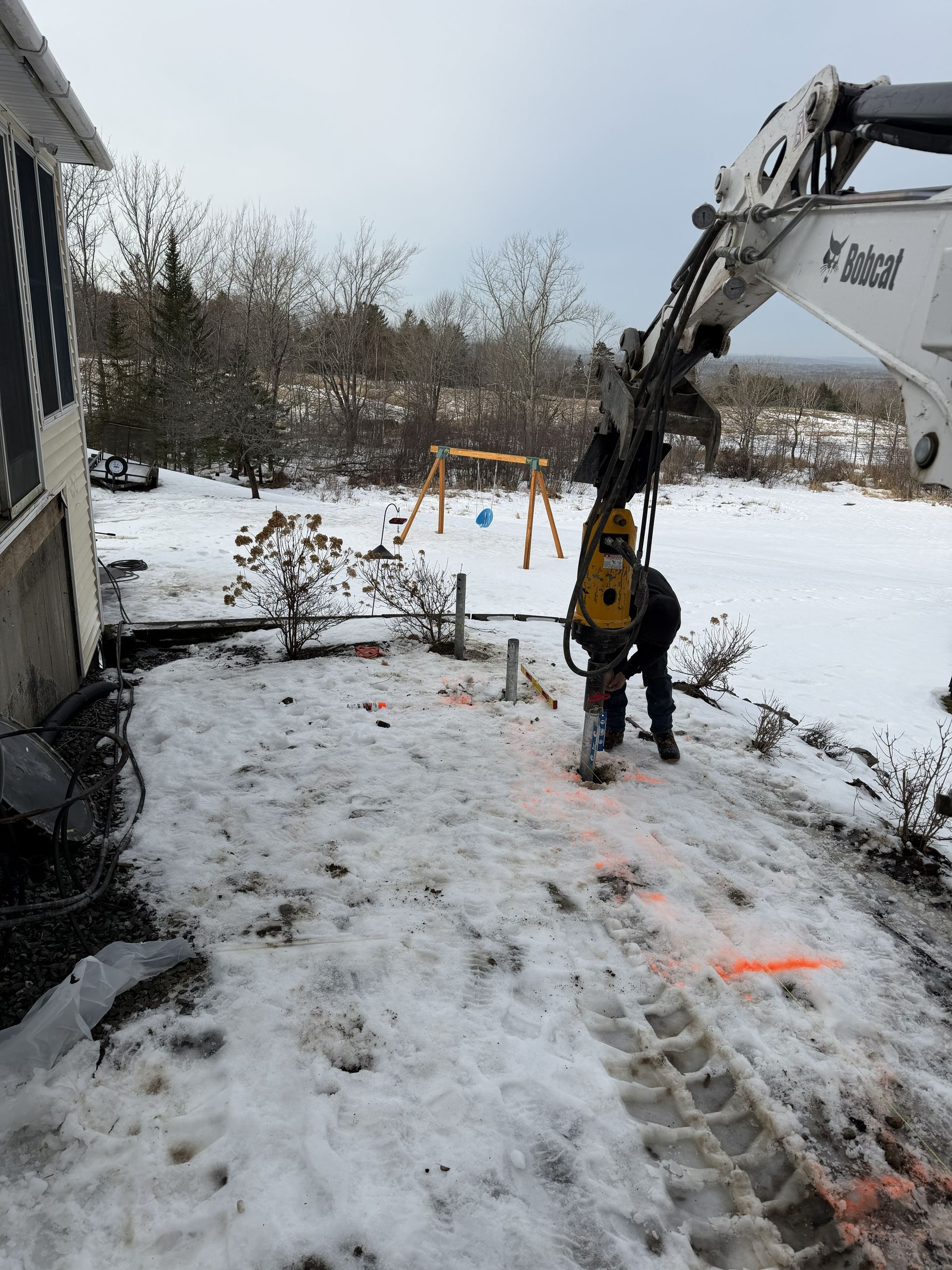 A person uses an excavator with a hydraulic pile driver to install metal poles into snowy ground near a house.