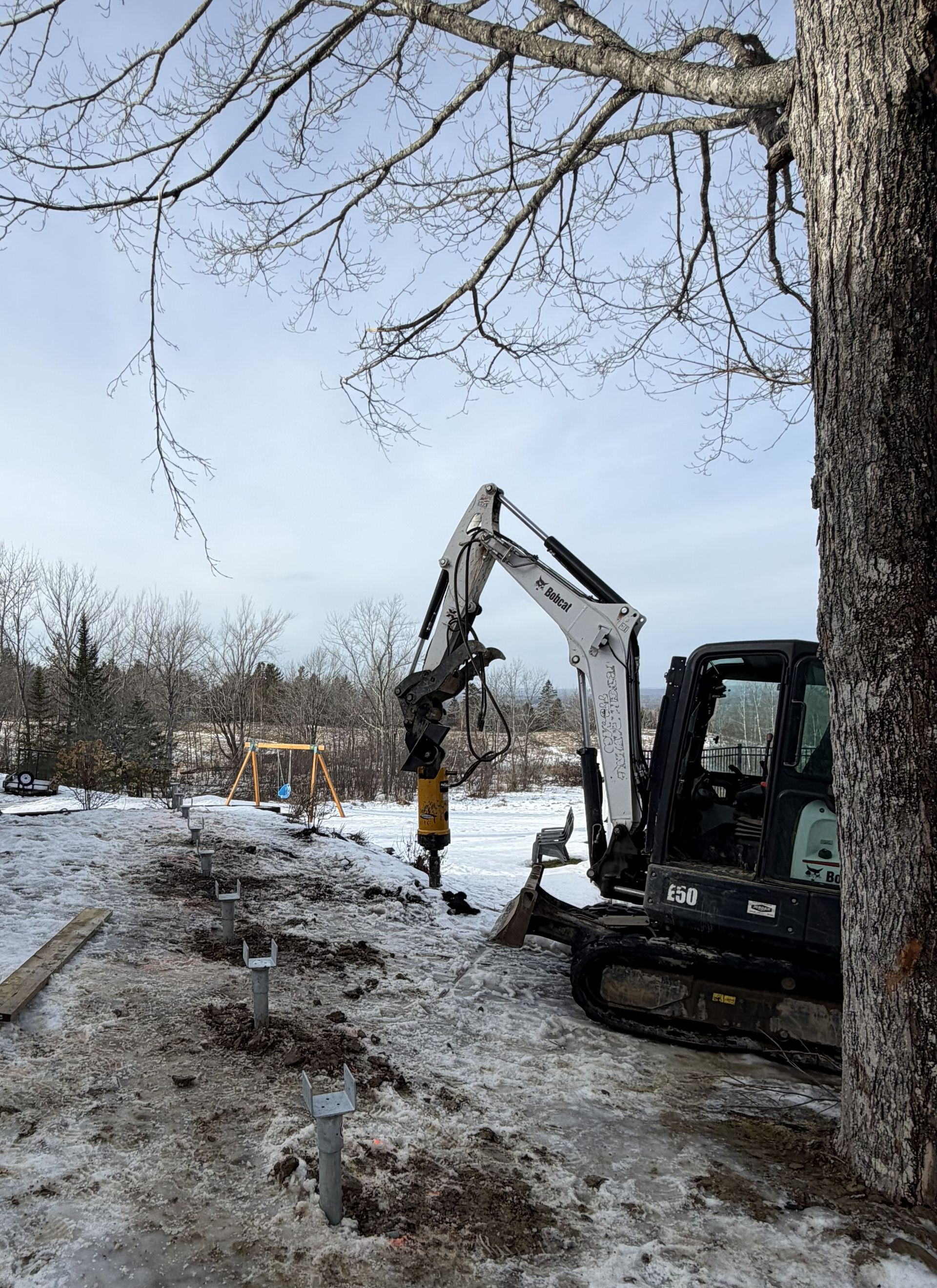 A small excavator with a hydraulic breaker attachment works on a snowy residential construction site with metal piers.