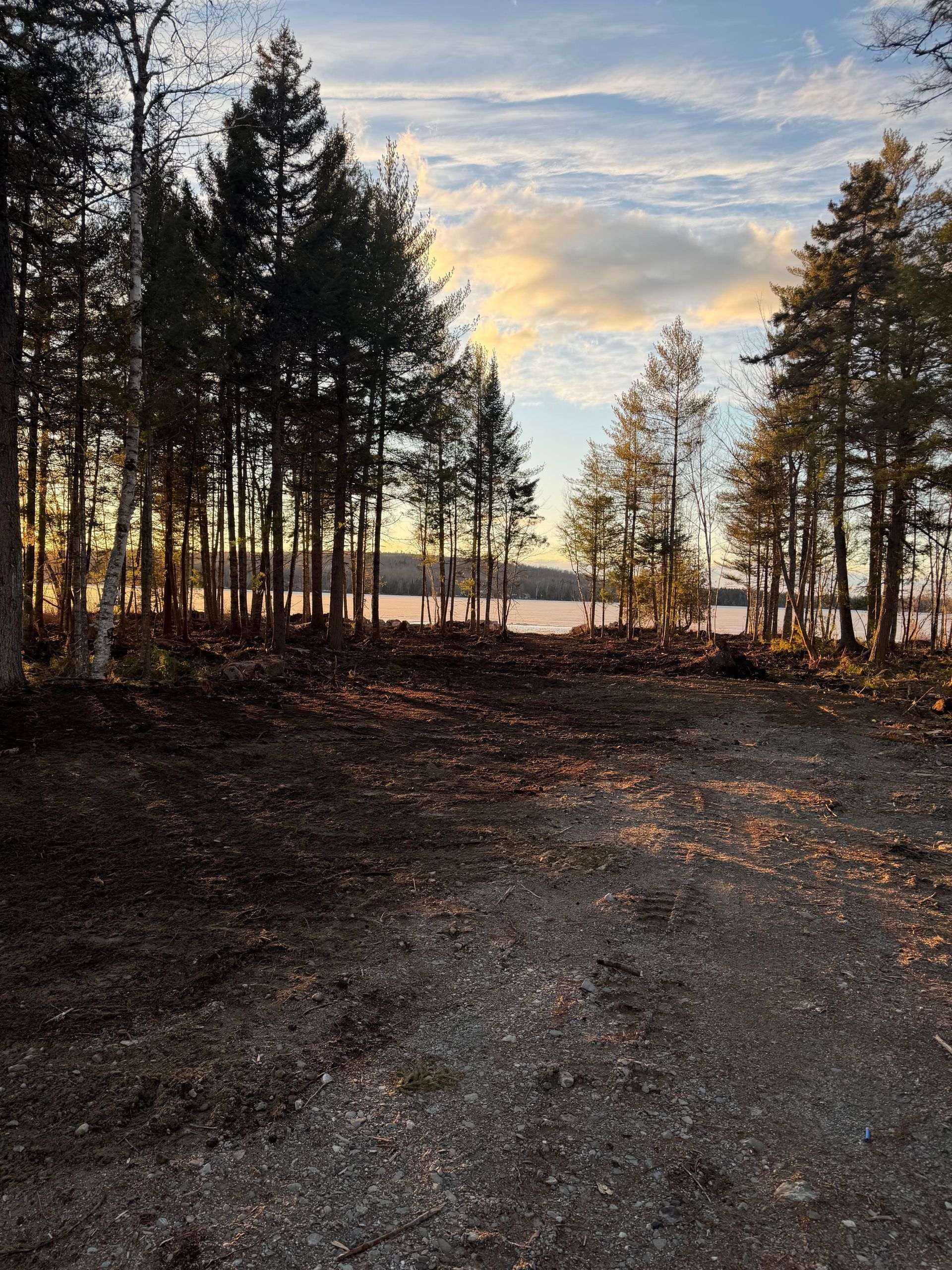An excavator with a hydraulic attachment works near a house on a dirt lot, with trees and a lake in the background.