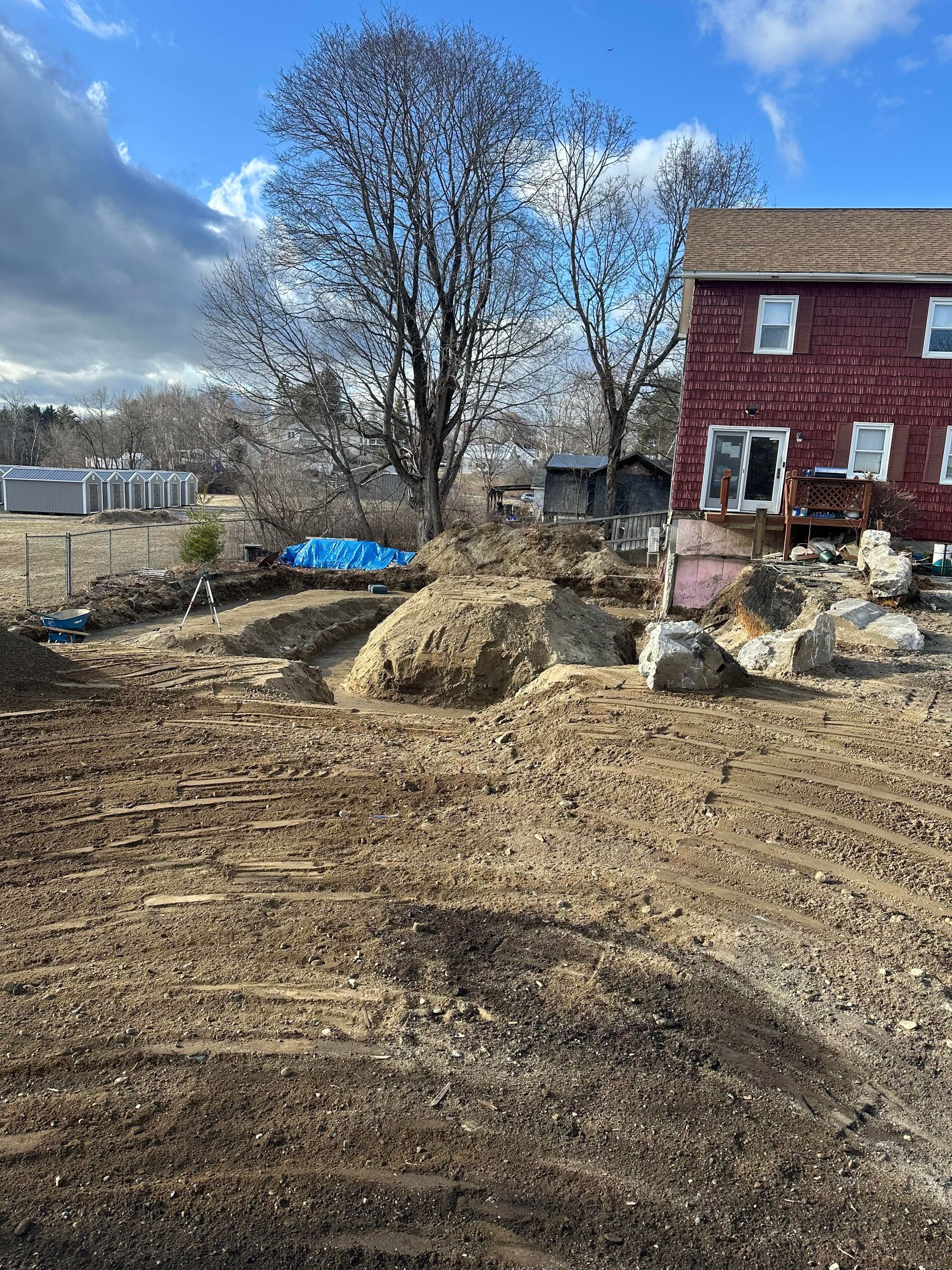A red house stands behind a yard with a dirt excavation, large rocks, and a blue tarp under bare trees on a sunny day.