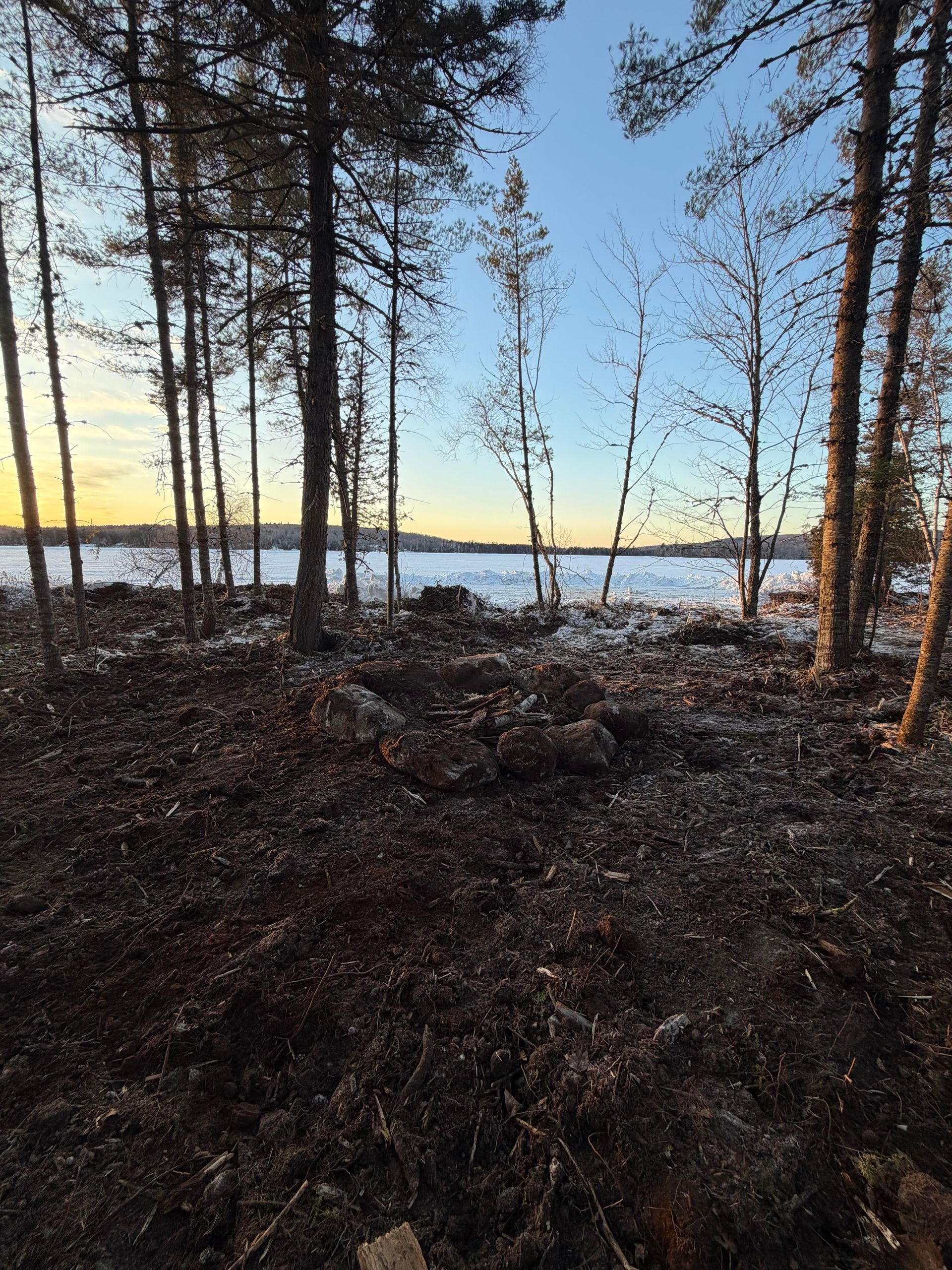 A fire pit sits in a forest clearing at dusk, overlooking a frozen, snow-dusted lake beneath a pale yellow and blue sky.
