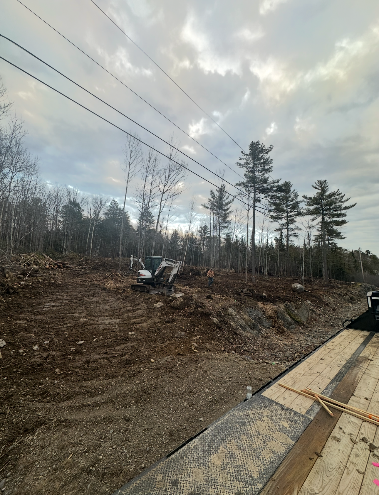 A white construction vehicle operates on a cleared, dirt-covered lot near a line of trees under a cloudy sky.