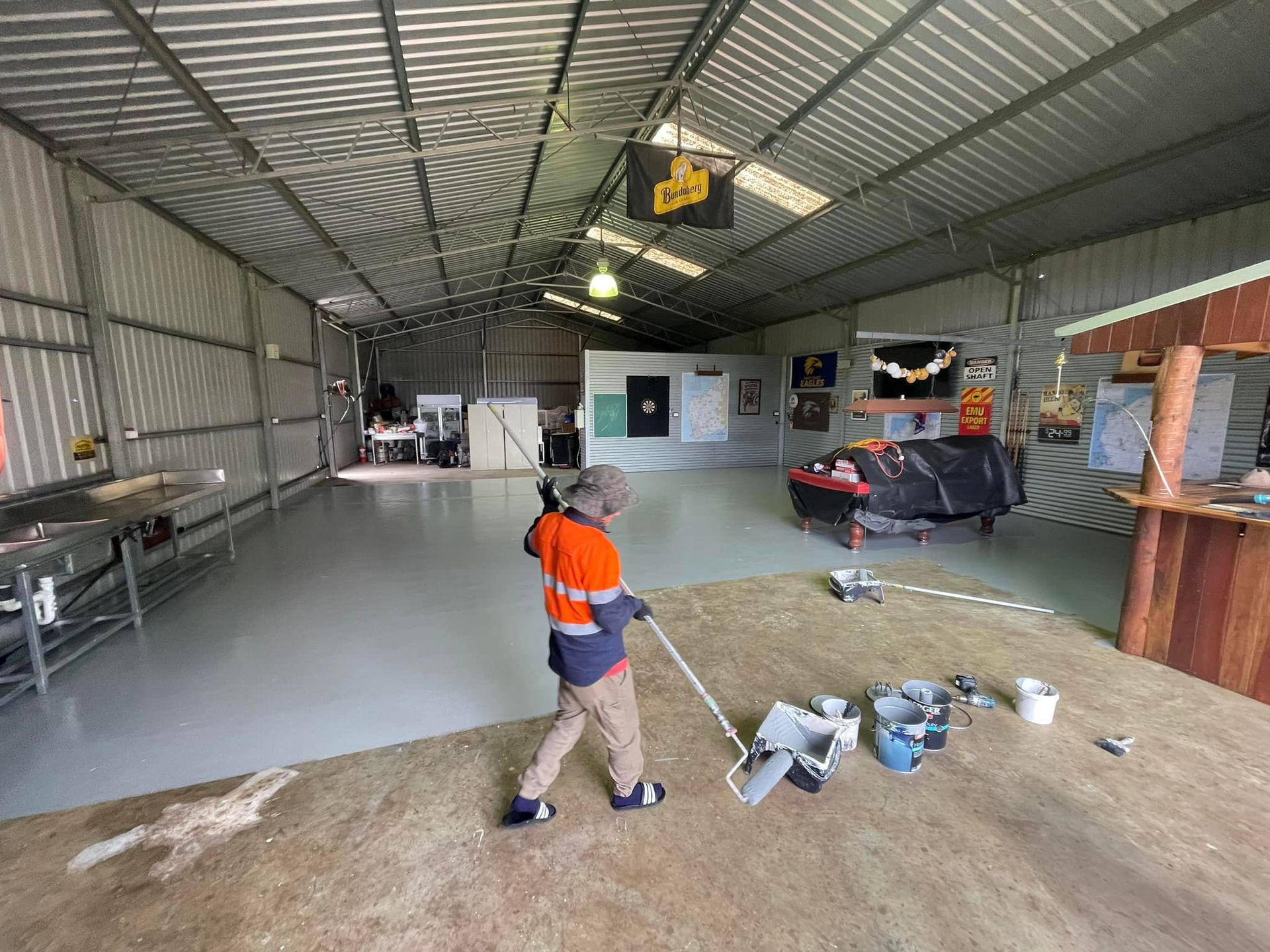 Person in work clothes sweeping a newly painted, light gray floor inside a metal shed.