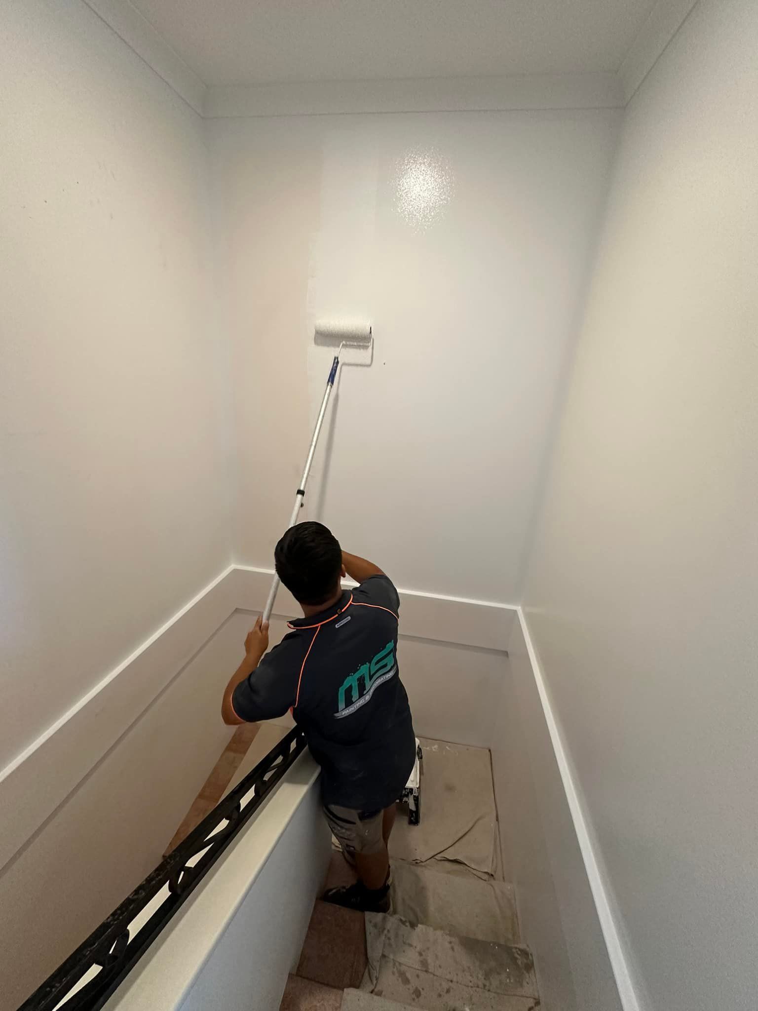 A person painting a white wall in a stairwell with a paint roller. They are wearing a dark shirt.