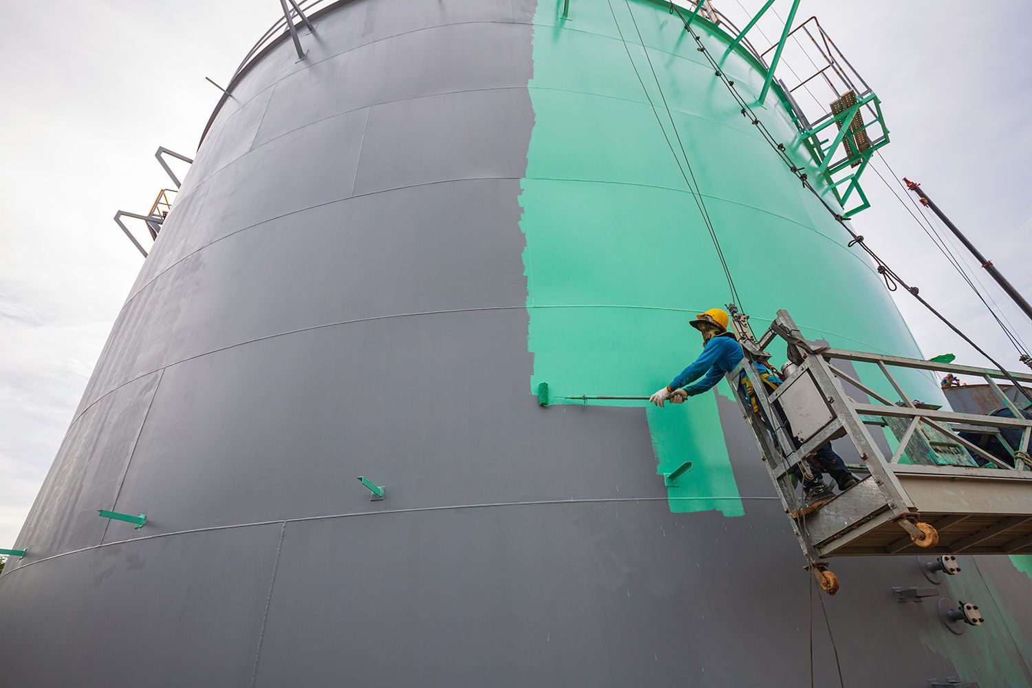 An industrial painter on a cable car uses a brush to paint the shell plate tank green. An industrial painter on a cable car uses a brush to paint the shell plate tank green.