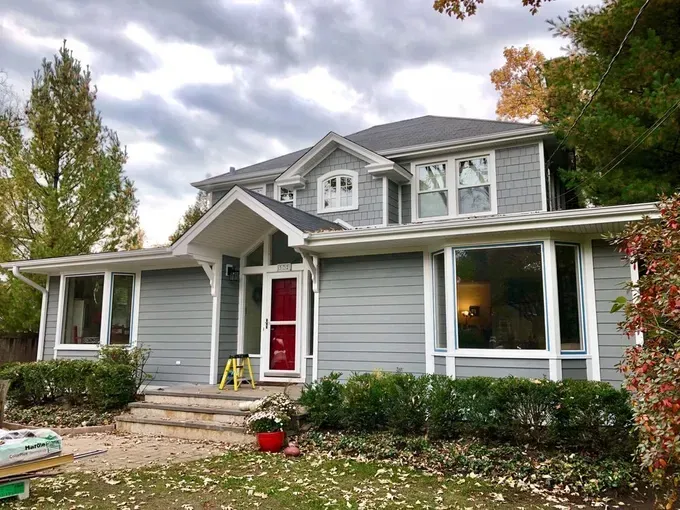 A gray house with white trim and a red door is being painted.