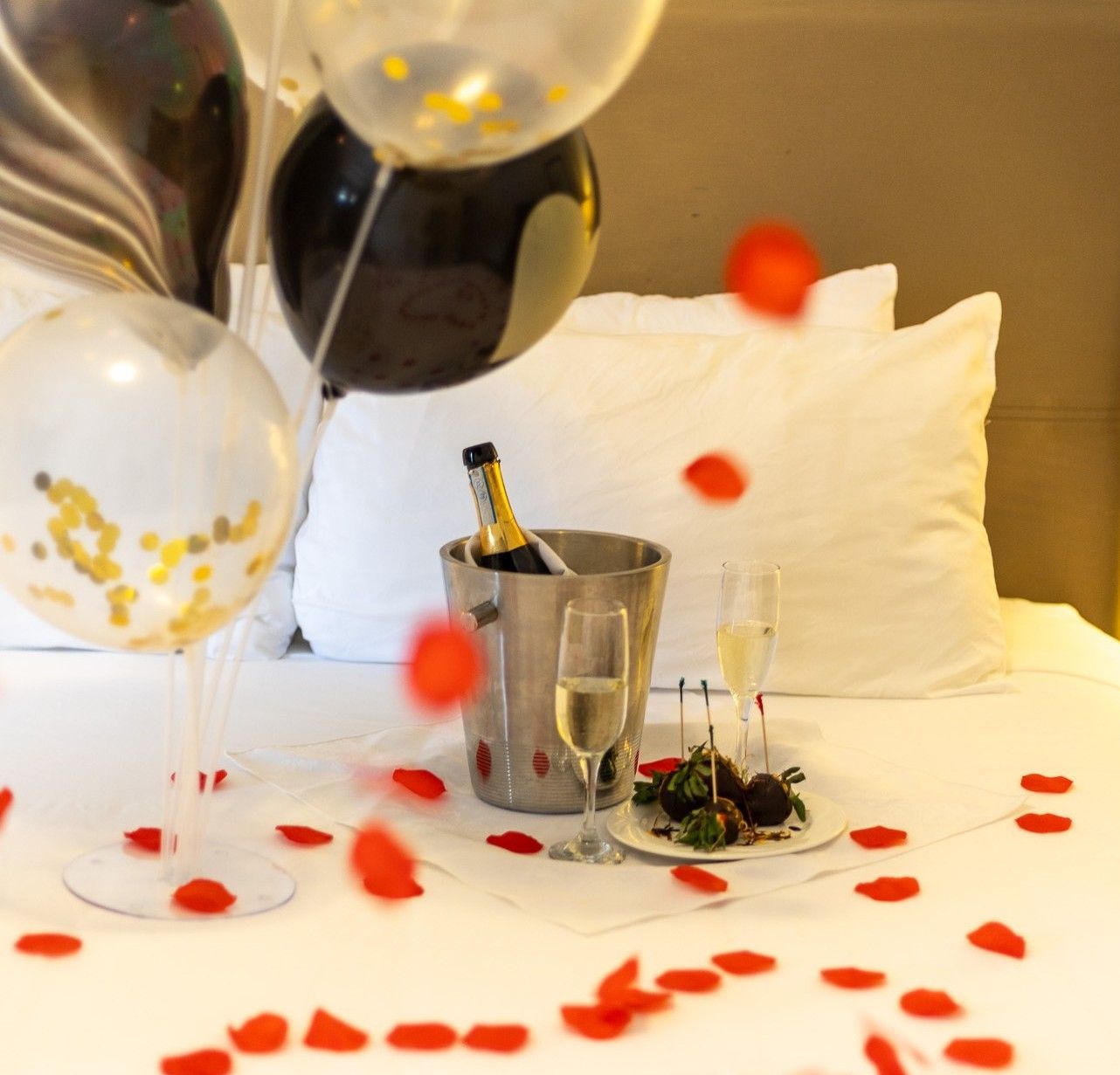 A bucket of champagne sits on a bed surrounded by balloons and rose petals