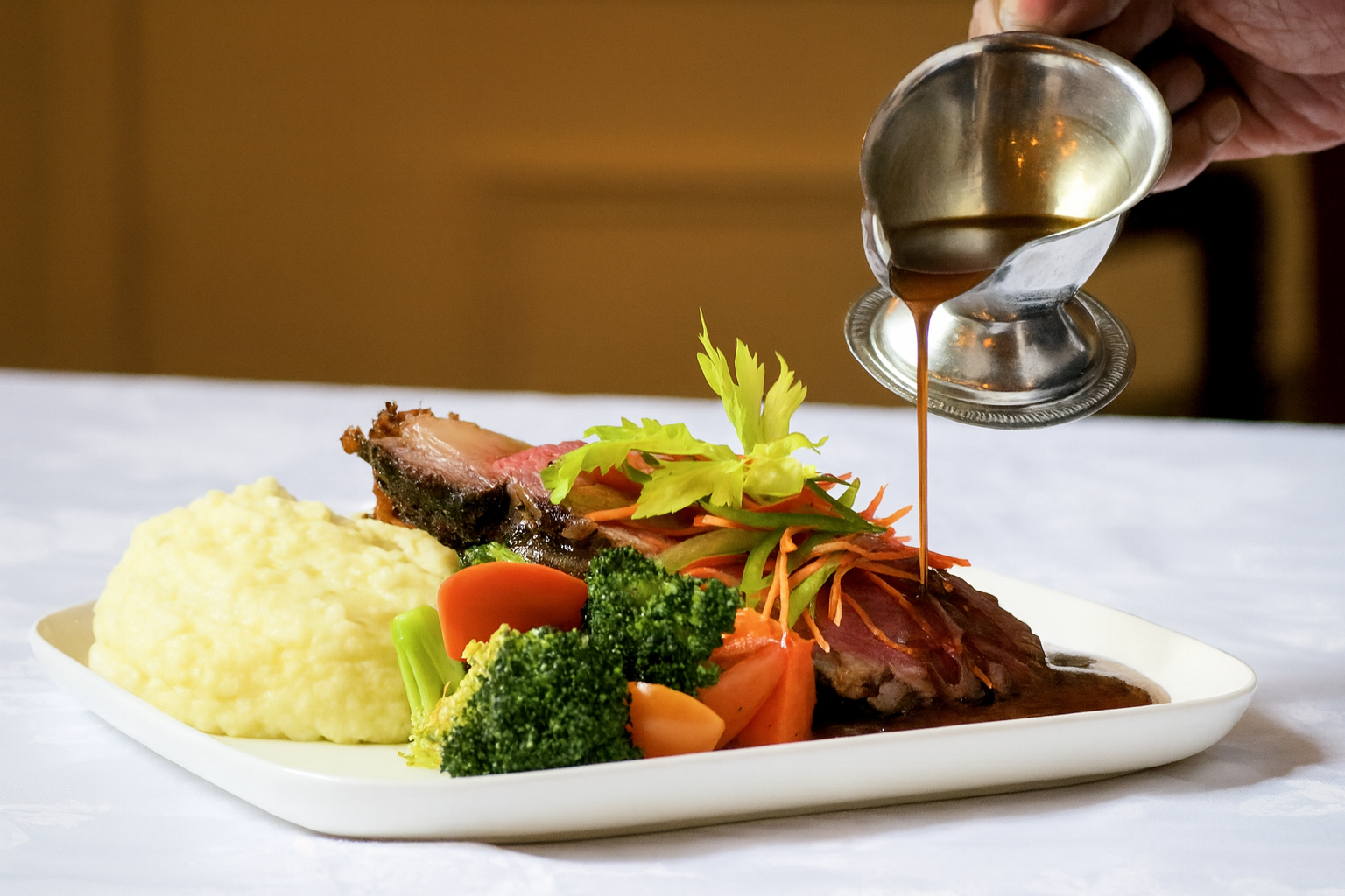 A plate of prime rib, vegetables, and mashed potatoes being drizzled with gravy.