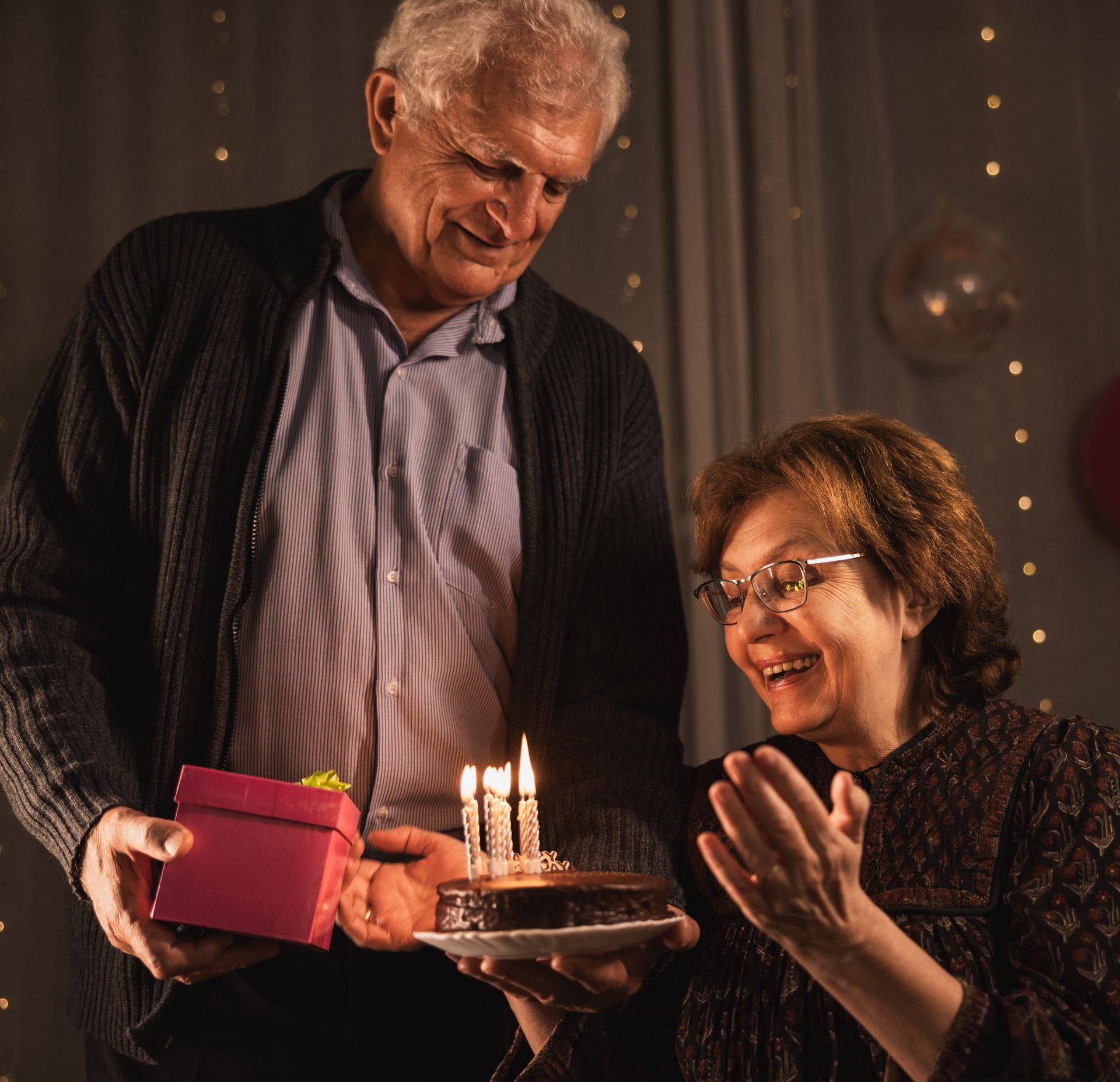 A man is giving a gift to a woman who is holding a birthday cake