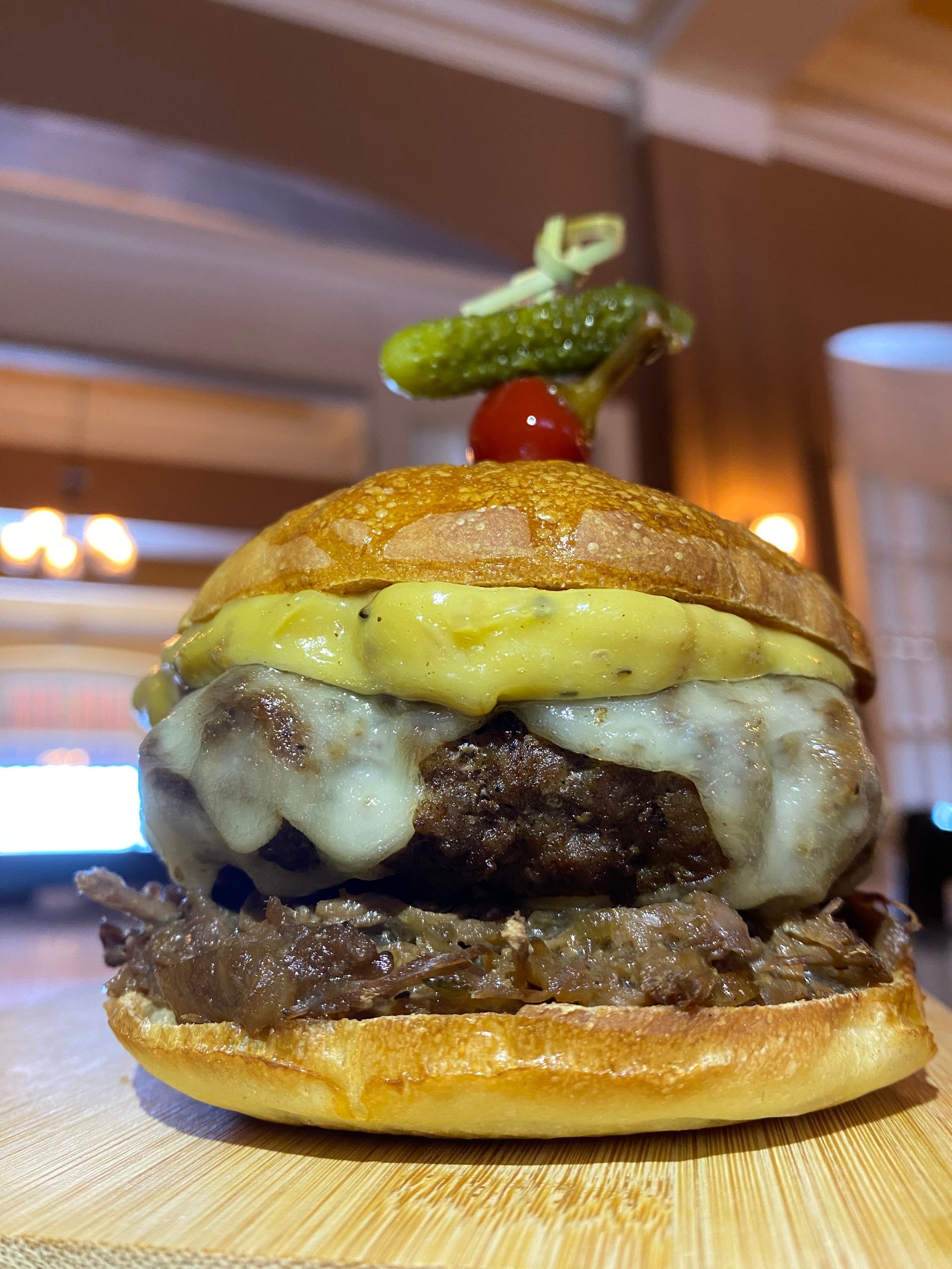 A gourmet burger with melted cheese and pulled meat on a bun, topped with a small pickle and cherry tomato on a skewer.