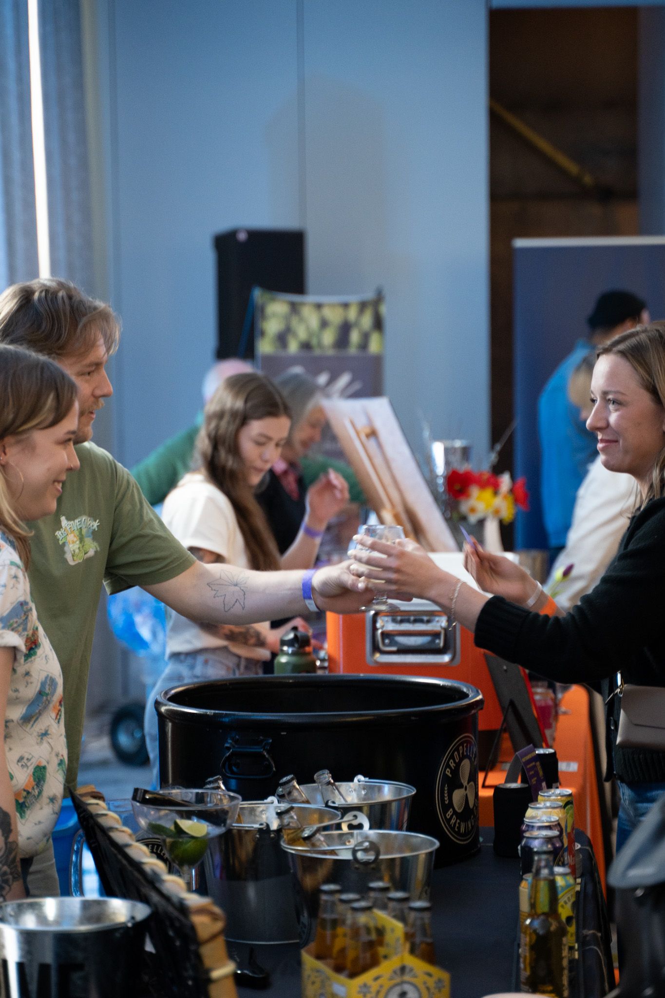 People at a food stall, exchanging items. A woman hands a cup to a man, smiling. Dark table, diverse items.
