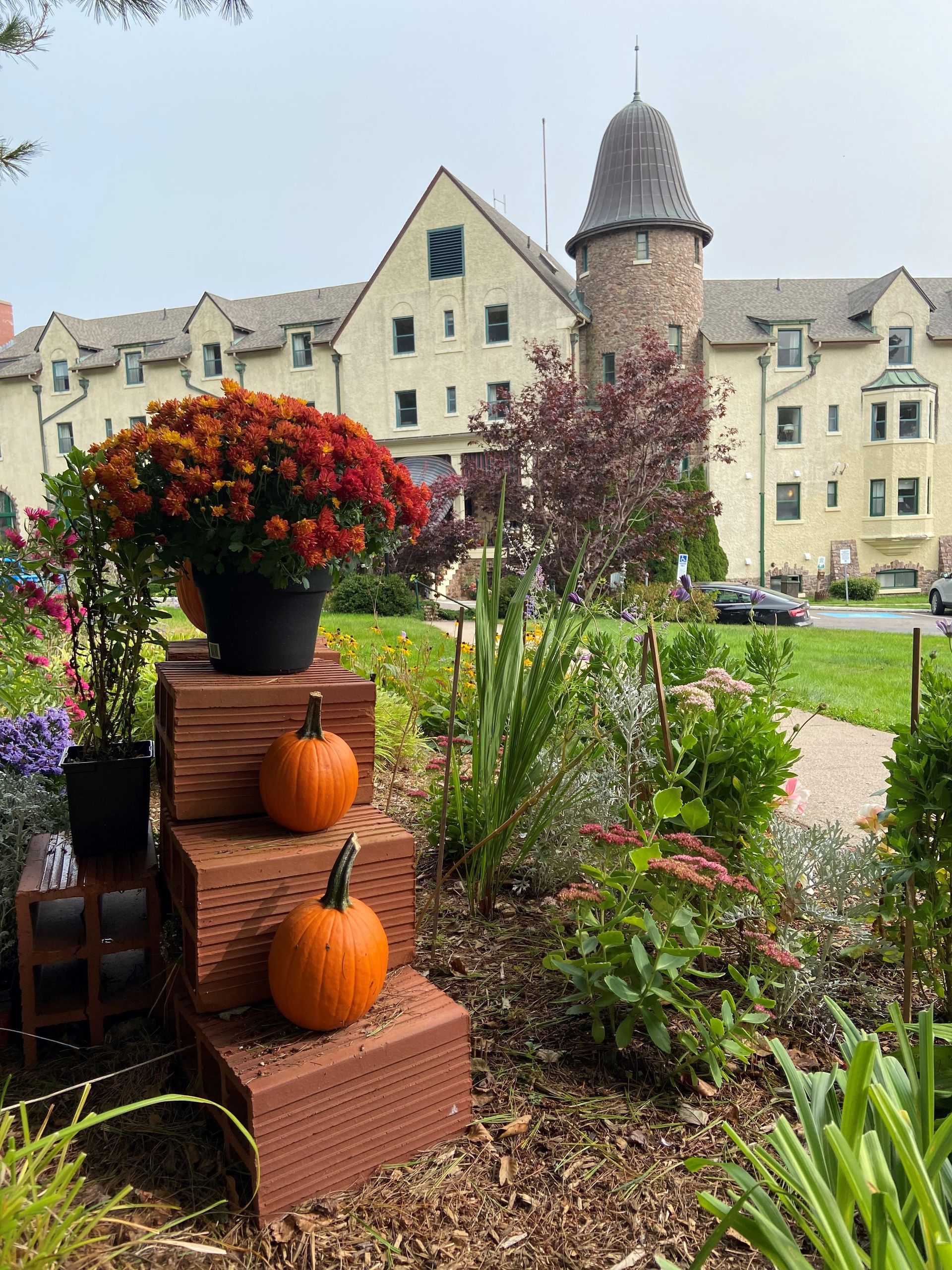 Autumn display with pumpkins, mums, and a stone tower in front of a light-colored building.