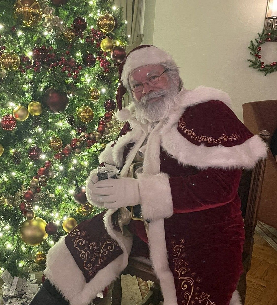 Santa Claus seated by a decorated Christmas tree, wearing a red suit and holding a gift.