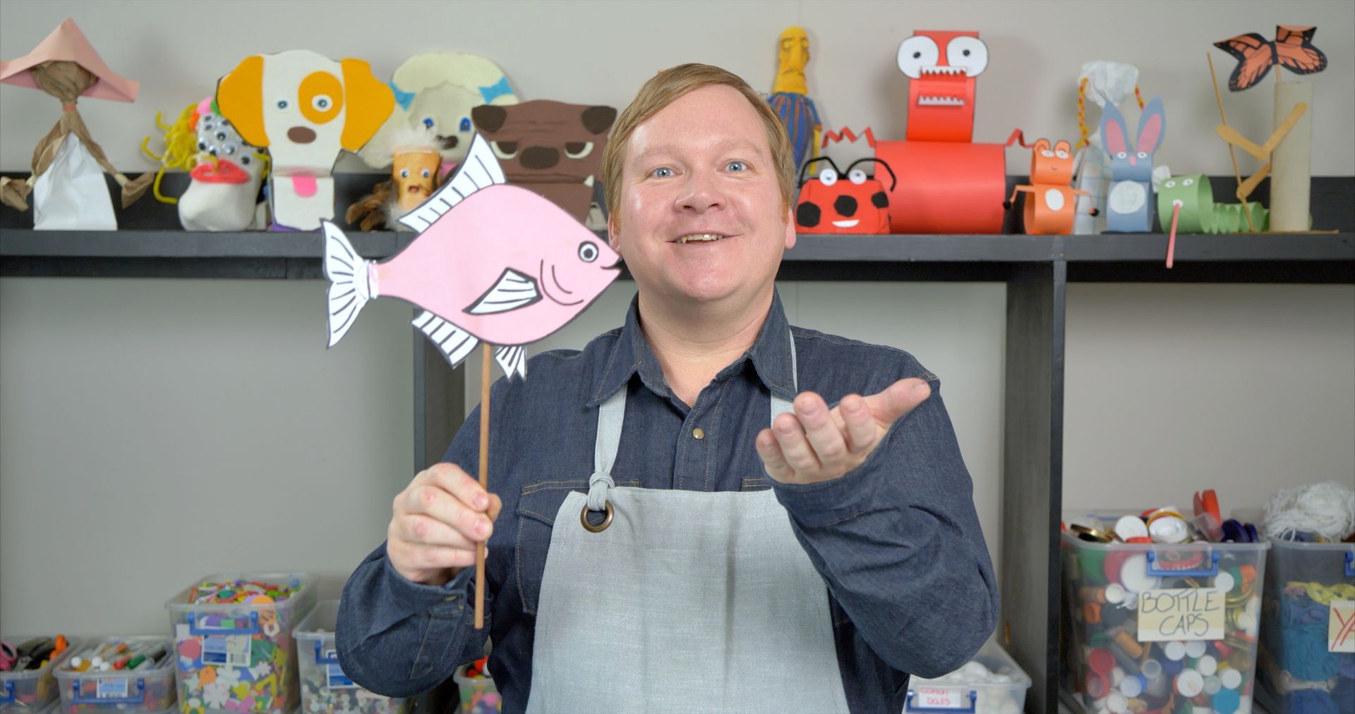 A man in an apron is holding a pink fish puppet.