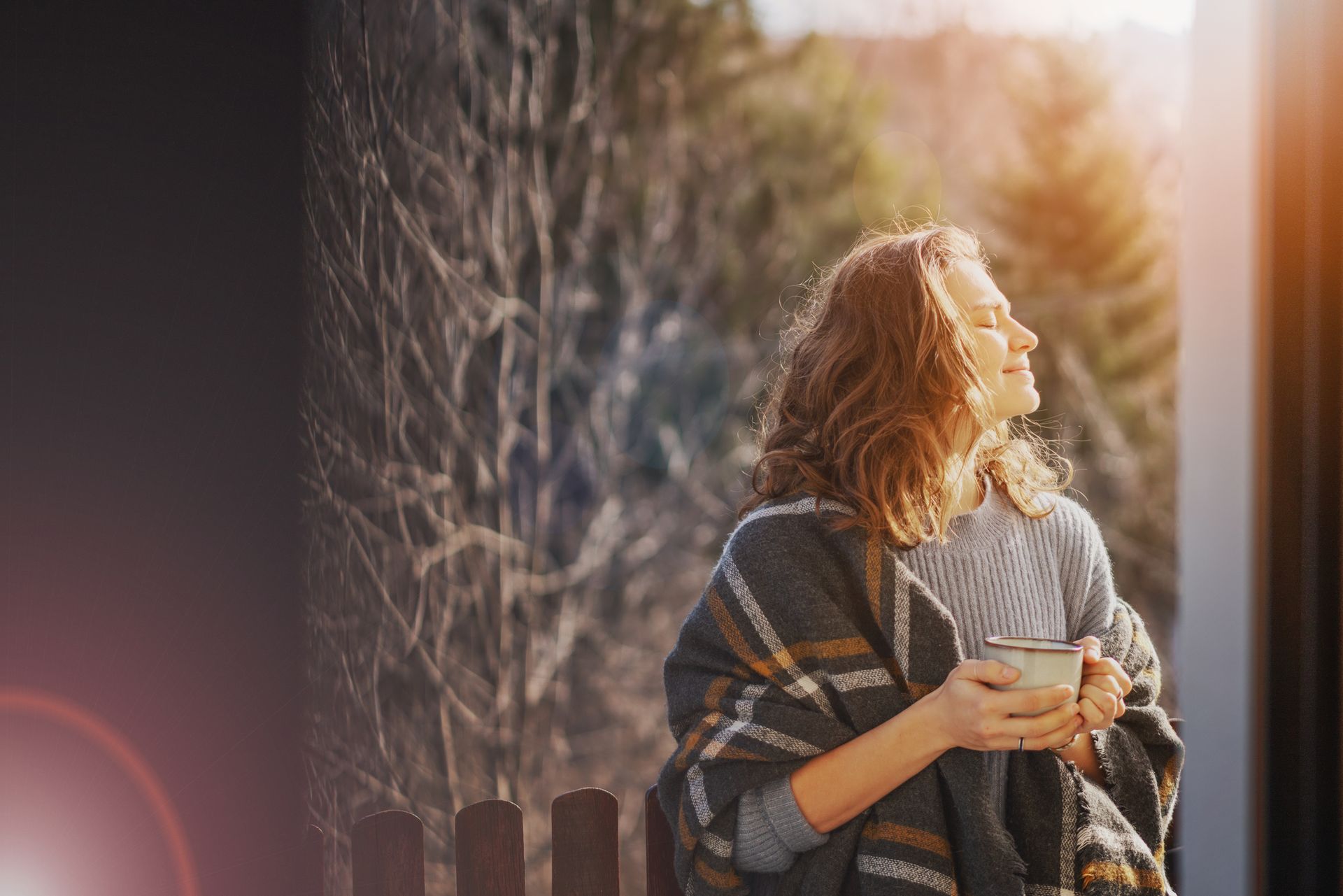 Woman in a shawl holding a mug, enjoying the sun, standing outside near a wooden fence.