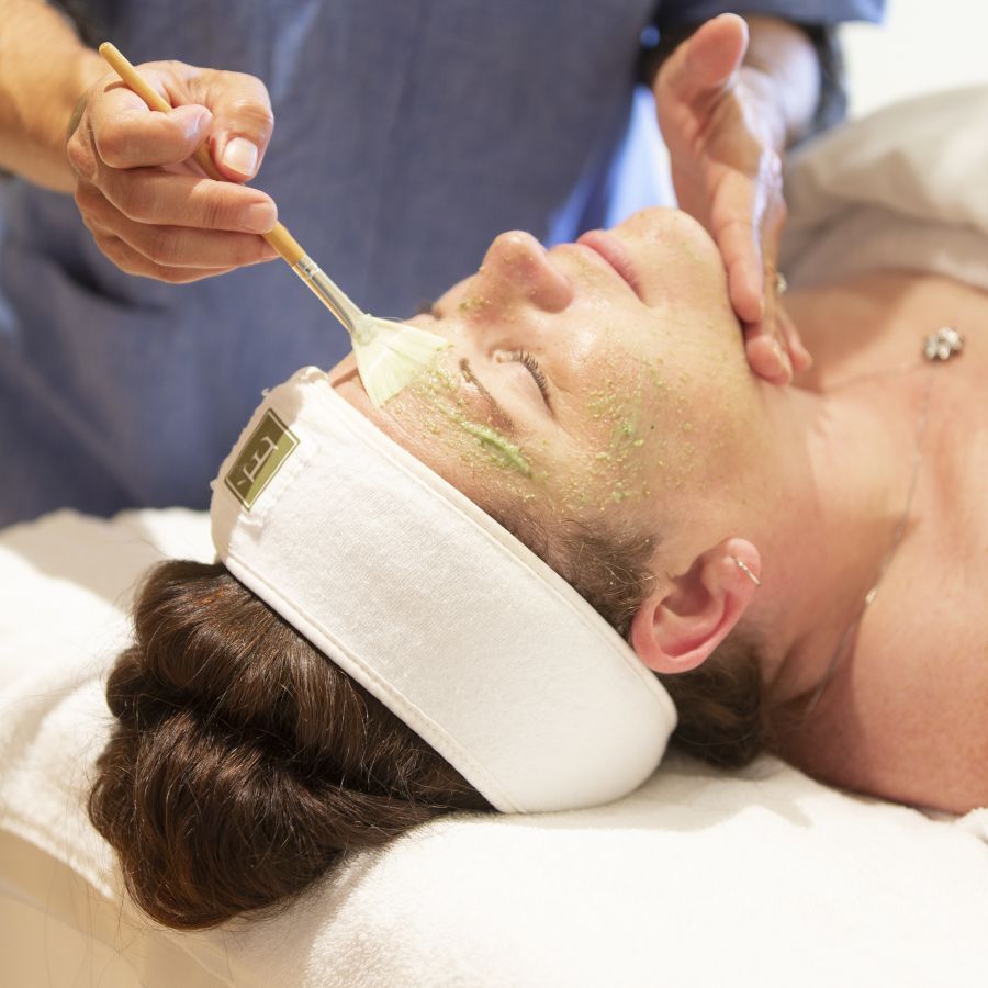 Person receiving a facial treatment at a spa. A practitioner applies a green mask with a brush.