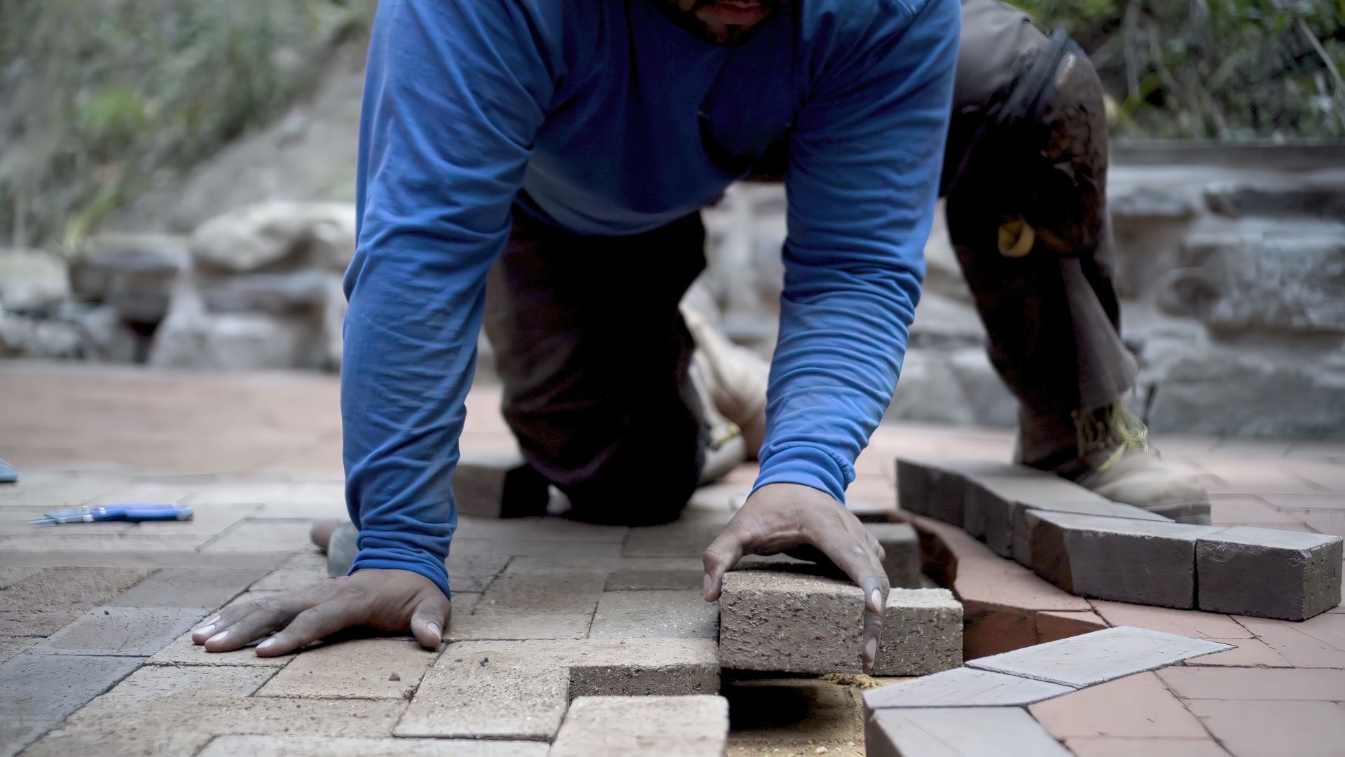 A man in a blue shirt is laying bricks on a sidewalk.