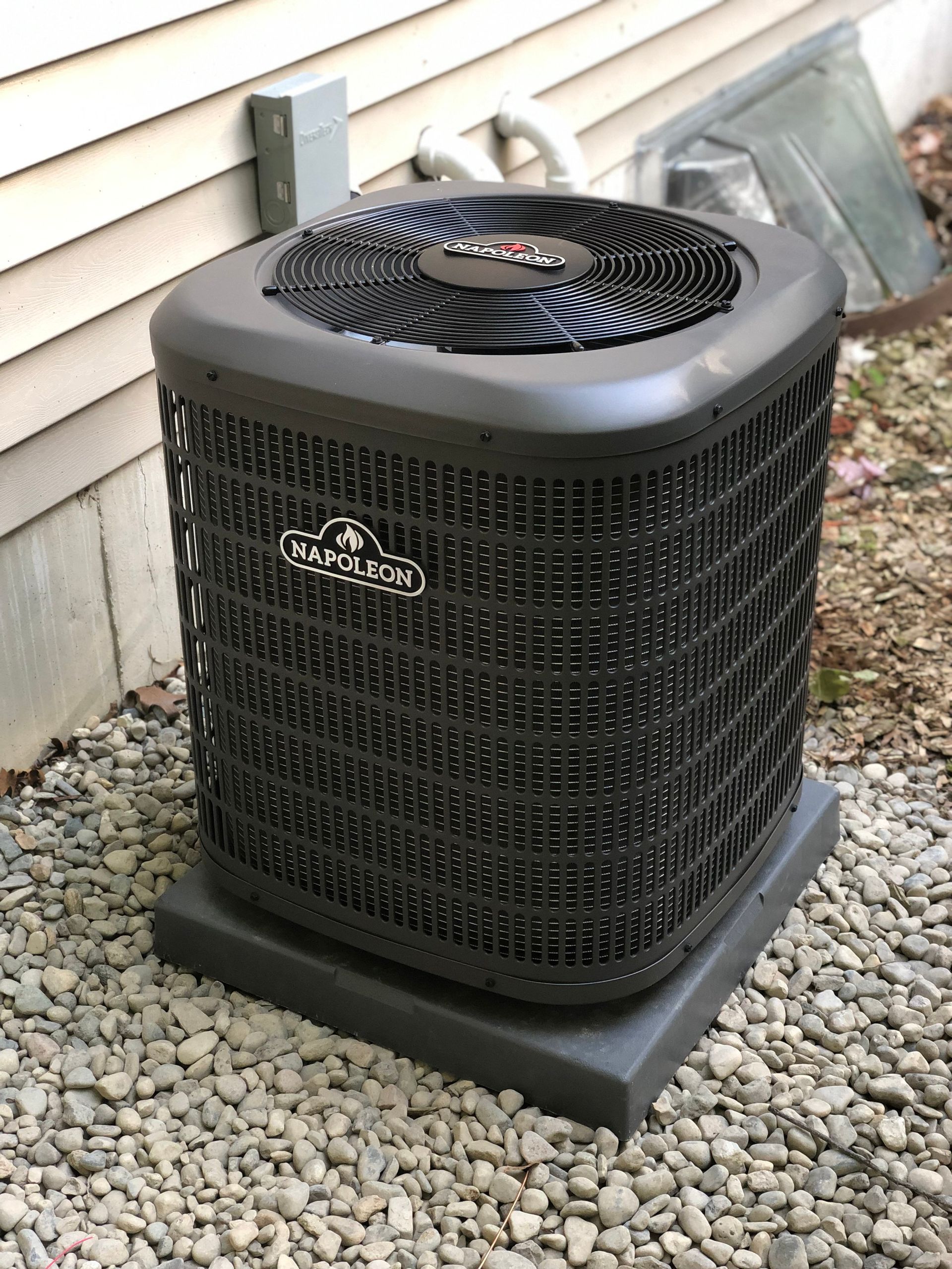 A black air conditioner is sitting on top of a pile of gravel next to a house.