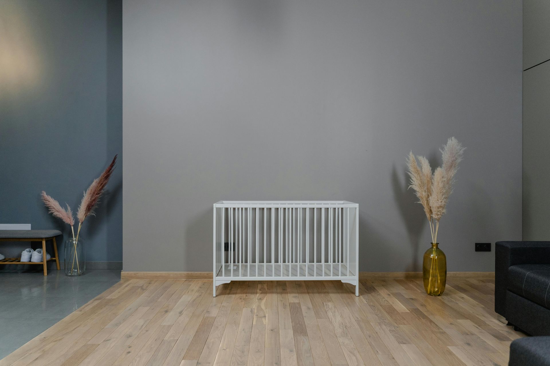 A white crib is sitting in a living room next to a vase of pampas grass.