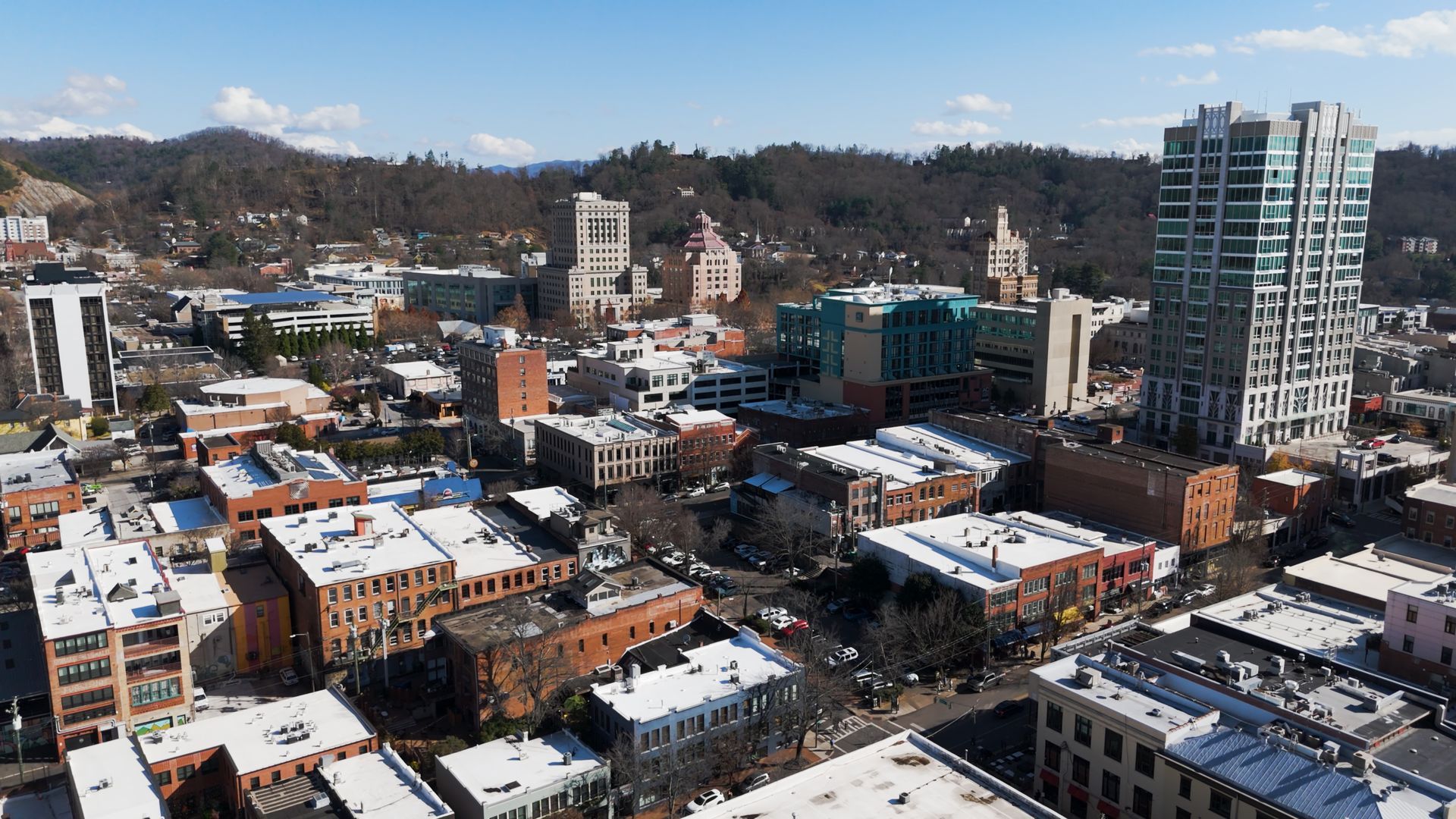 Aerial view of a city with buildings of various sizes and architectural styles, mountains in the background under a blue sky.