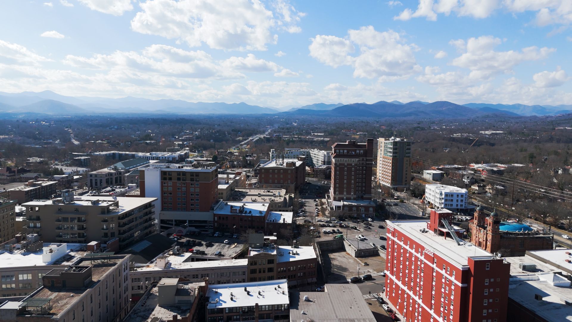 Aerial view of a city with tall buildings, streets, and mountains in the background under a blue sky.
