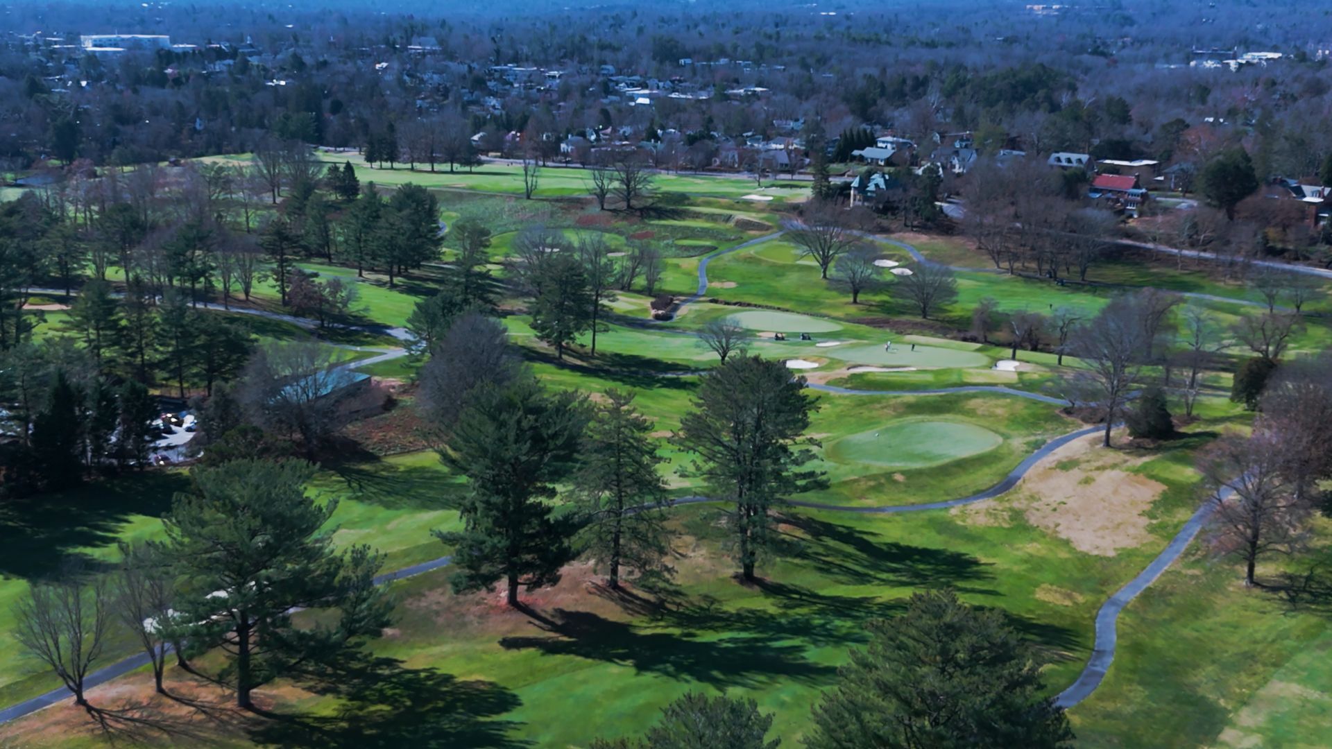 Aerial view of a green golf course with trees, fairways, and a distant town, under a clear sky.