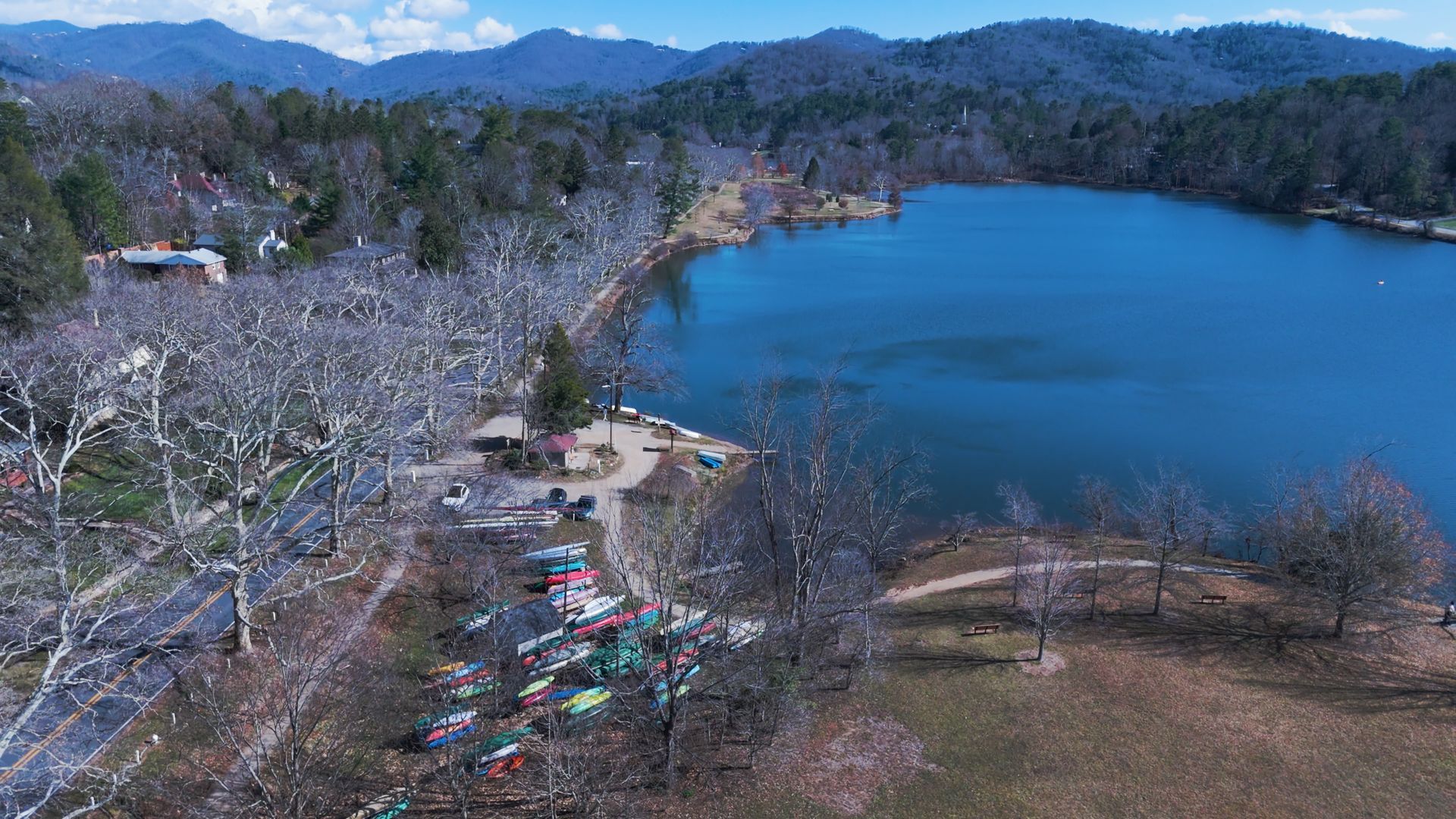 Lake scene with trees, park, and mountains in the background under a blue sky.