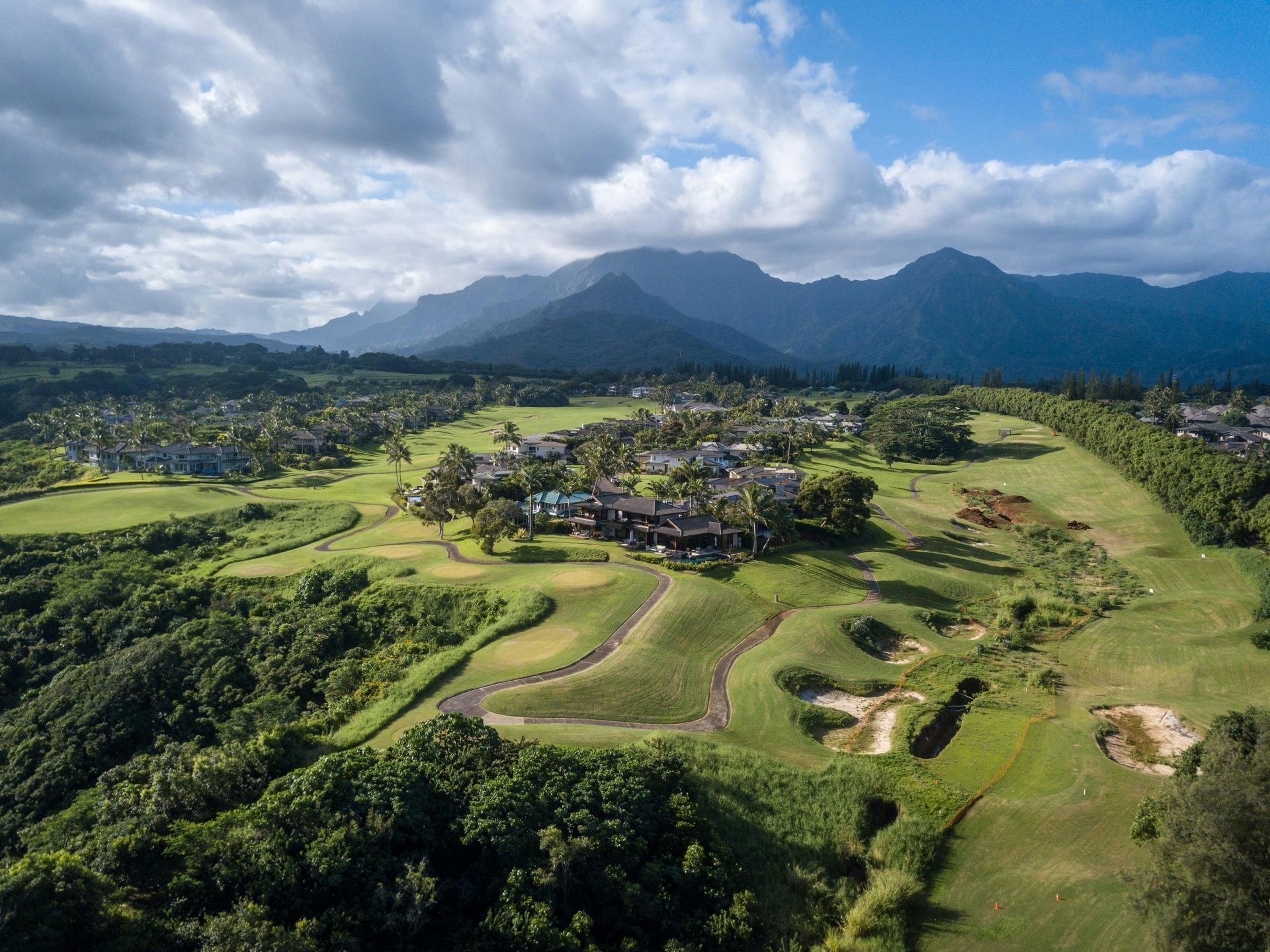 Aerial view of a lush green golf course with a resort complex, mountains, and partly cloudy sky.