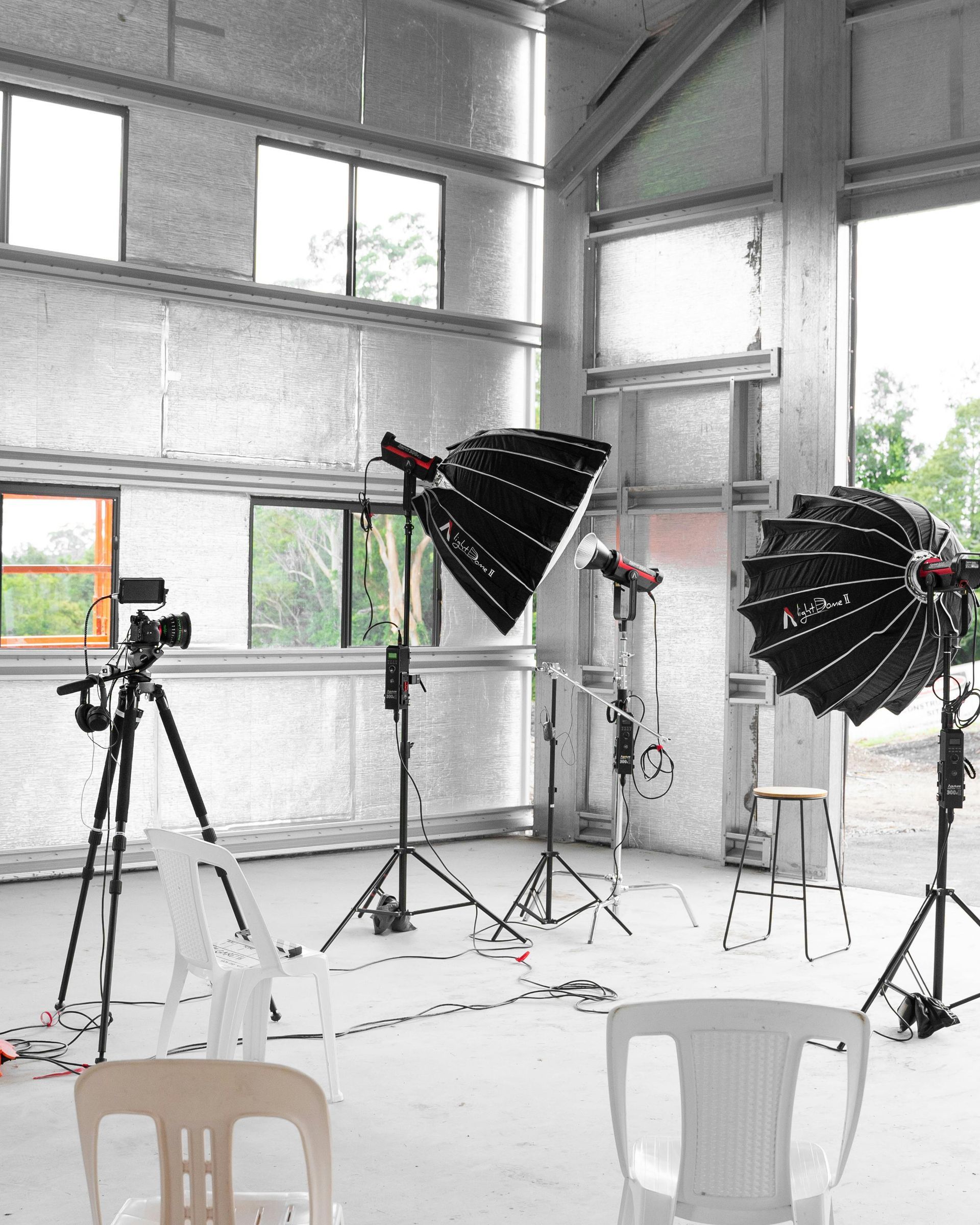 A film set inside a studio under construction, featuring a camera on a tripod, two large studio lights, and three chairs.